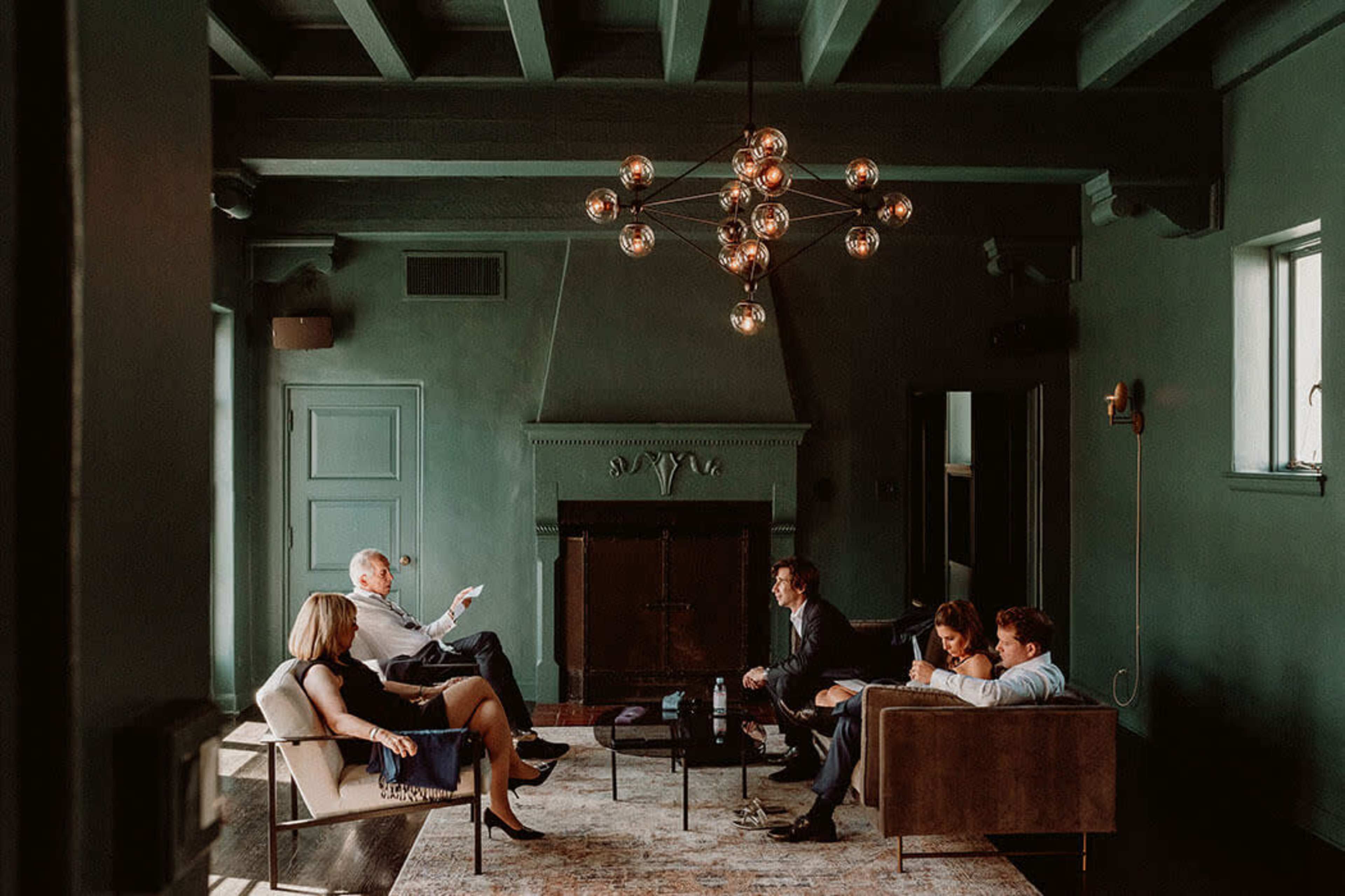 A group of six people sits in a dimly lit room with green walls, engaged in conversation around a low coffee table.