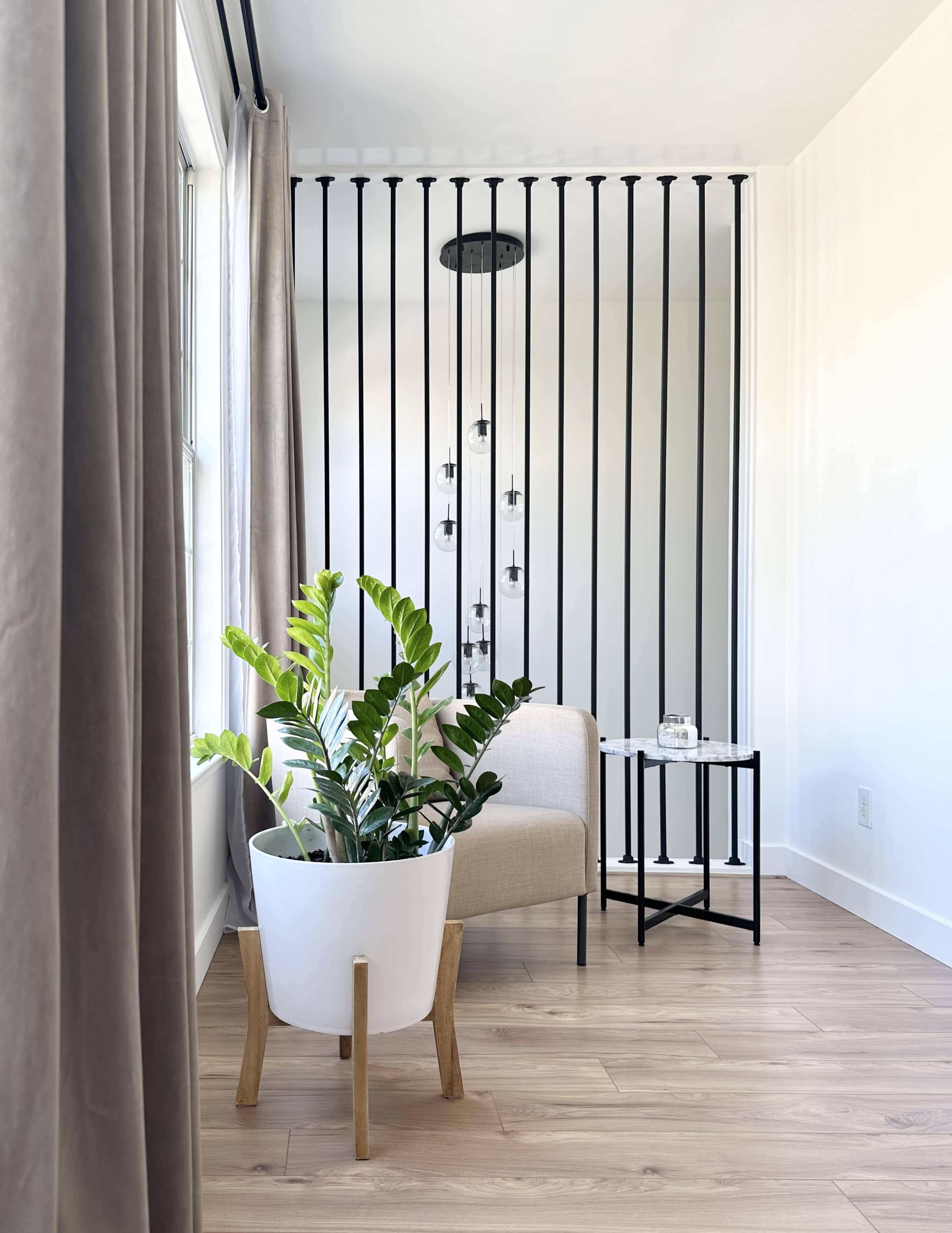 A light-filled interior features a beige chair next to a small black table, with a large potted plant and a decorative item on the table, separated by vertical black bars in the background.