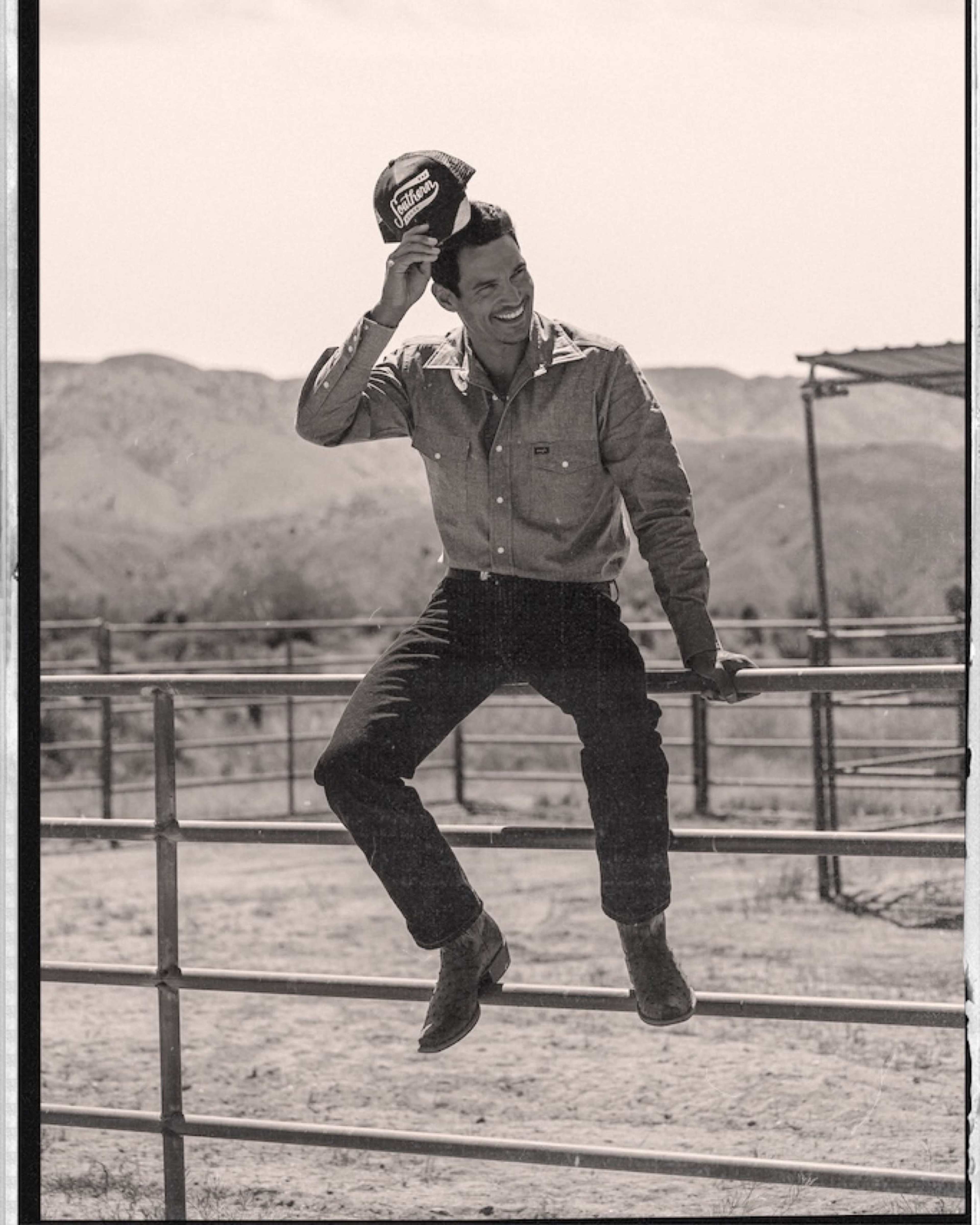 A man wearing a denim shirt and cowboy hat sits on a fence with mountains in the background.
