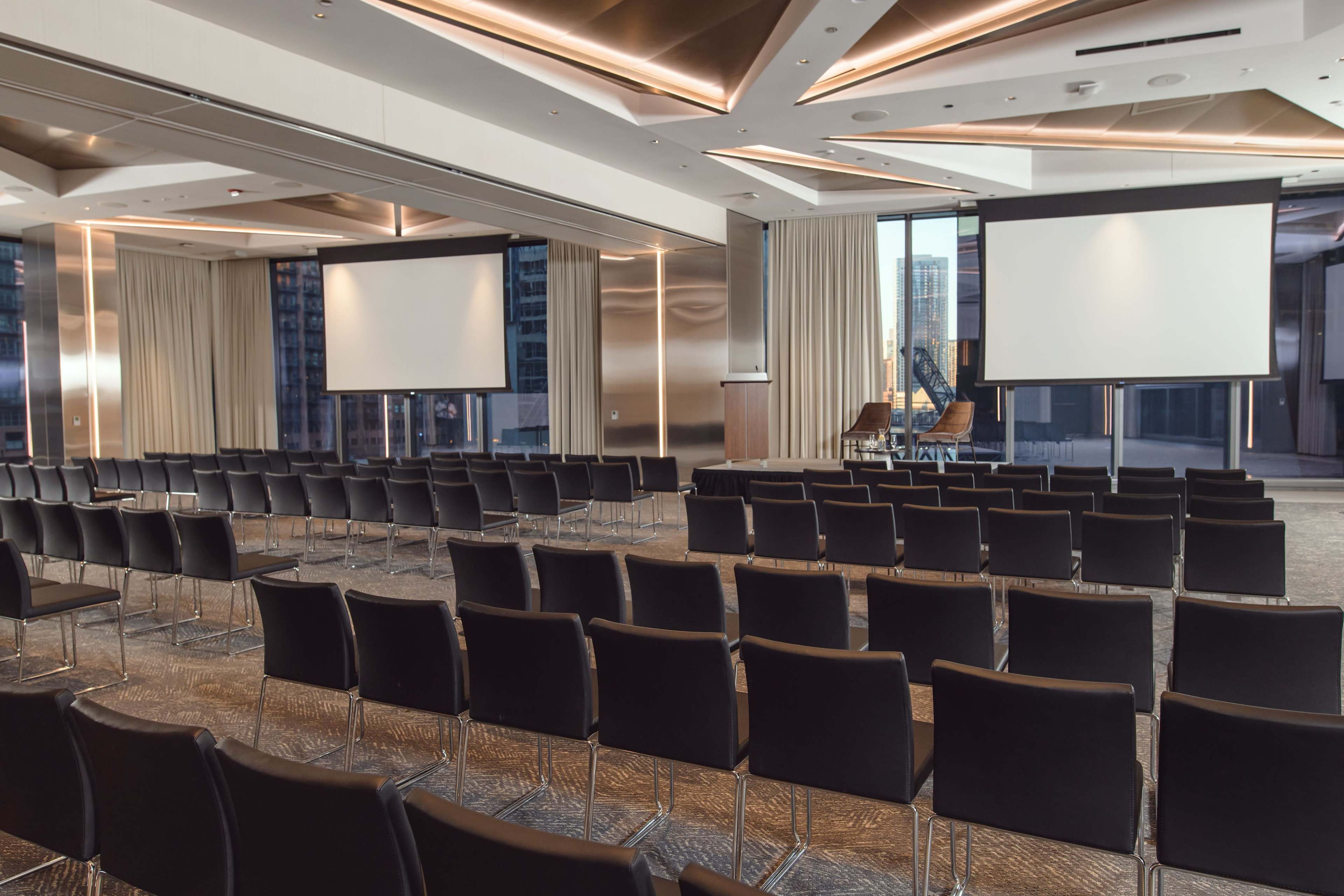 The image shows a modern conference room set up with rows of black chairs facing two large screens and a speaker's podium, flanked by large windows overlooking a cityscape.