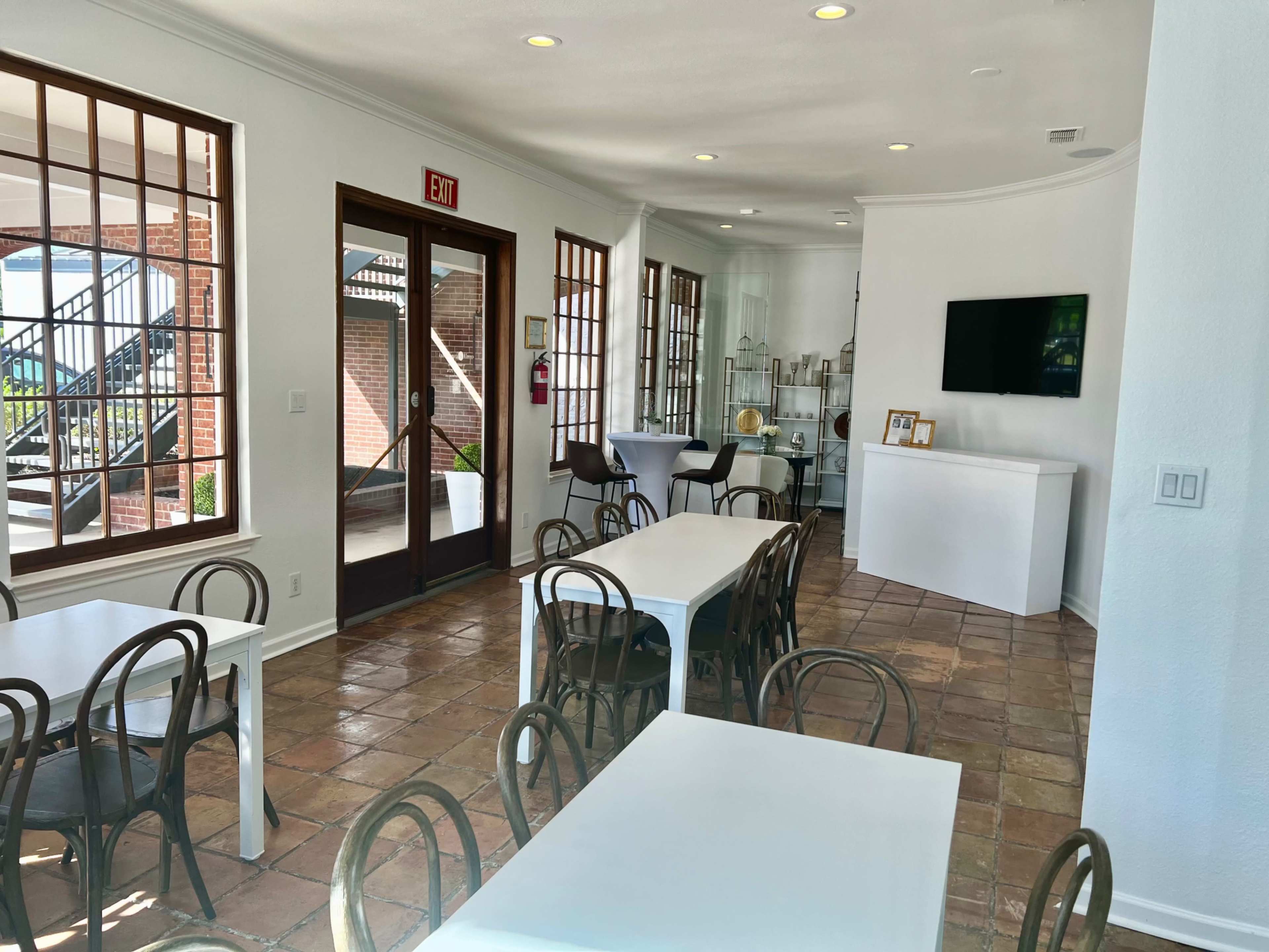 A bright, open interior space with several white tables, a television on the wall, and large windows providing a view of the exterior.