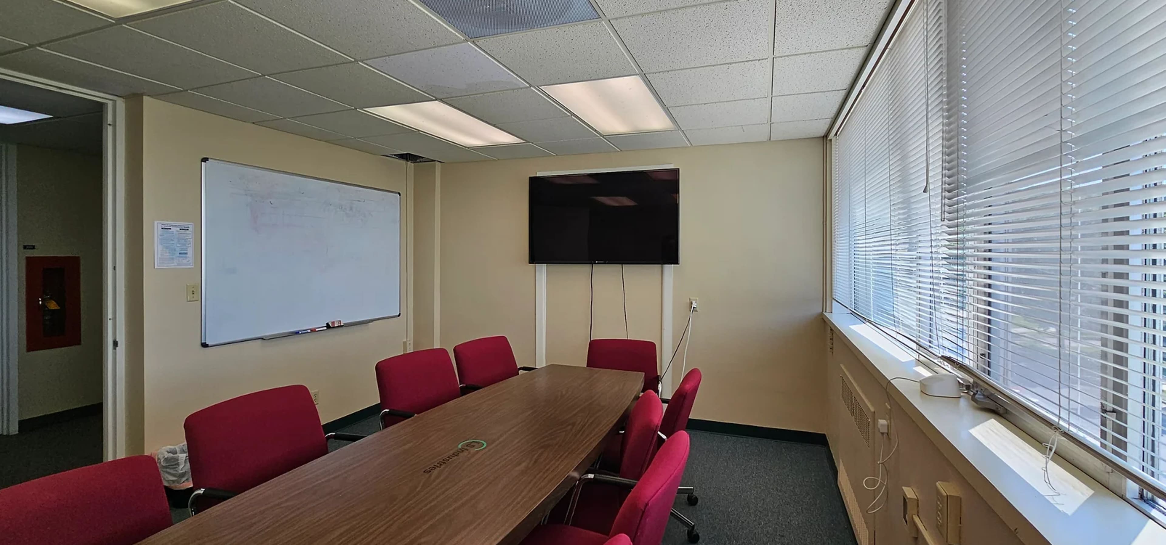 A conference room with a long wooden table surrounded by red chairs, a whiteboard on one wall, and a mounted television on another wall.