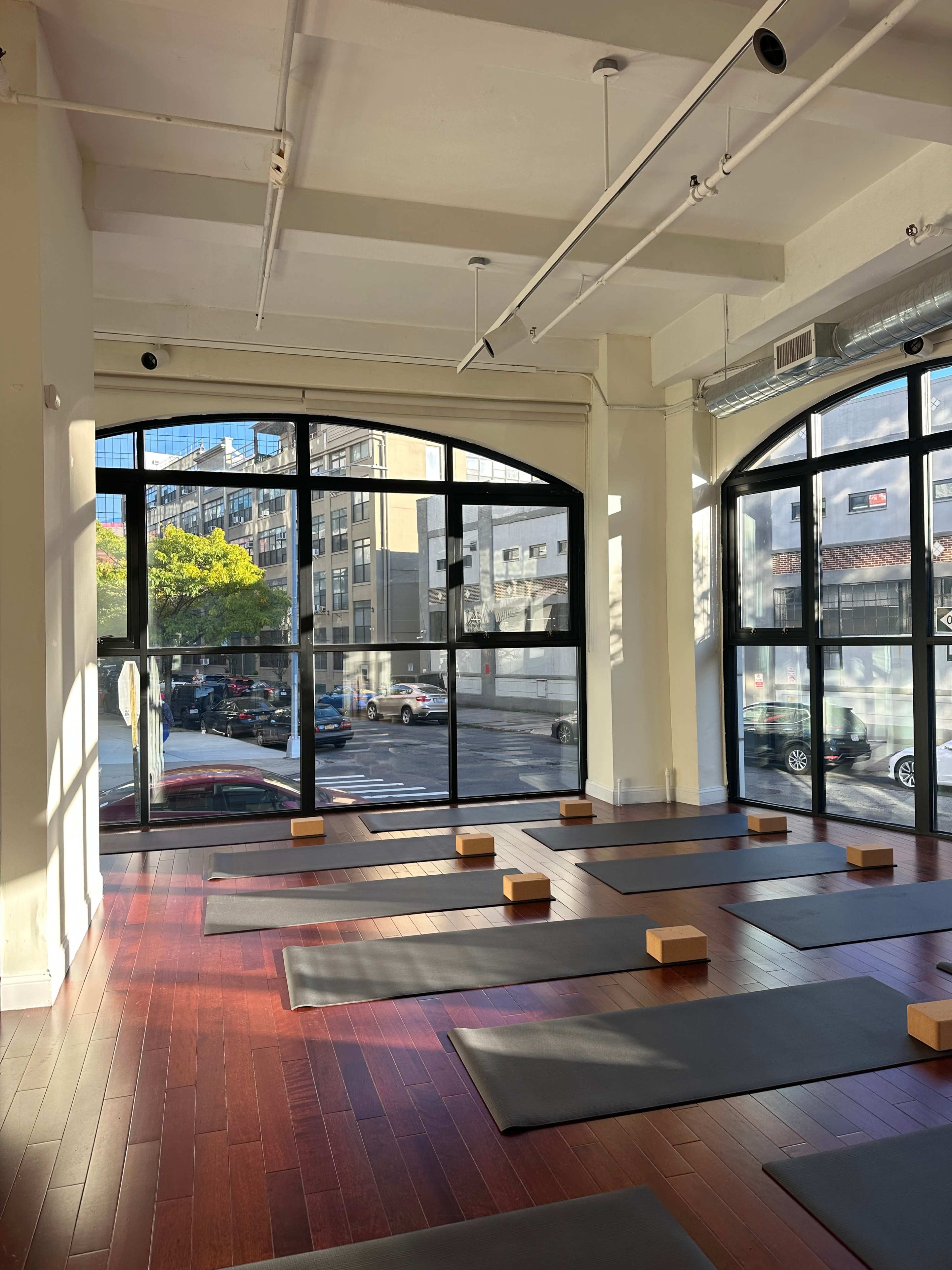 A yoga studio with several mats and wooden blocks arranged on a polished wooden floor, illuminated by natural light from large windows.