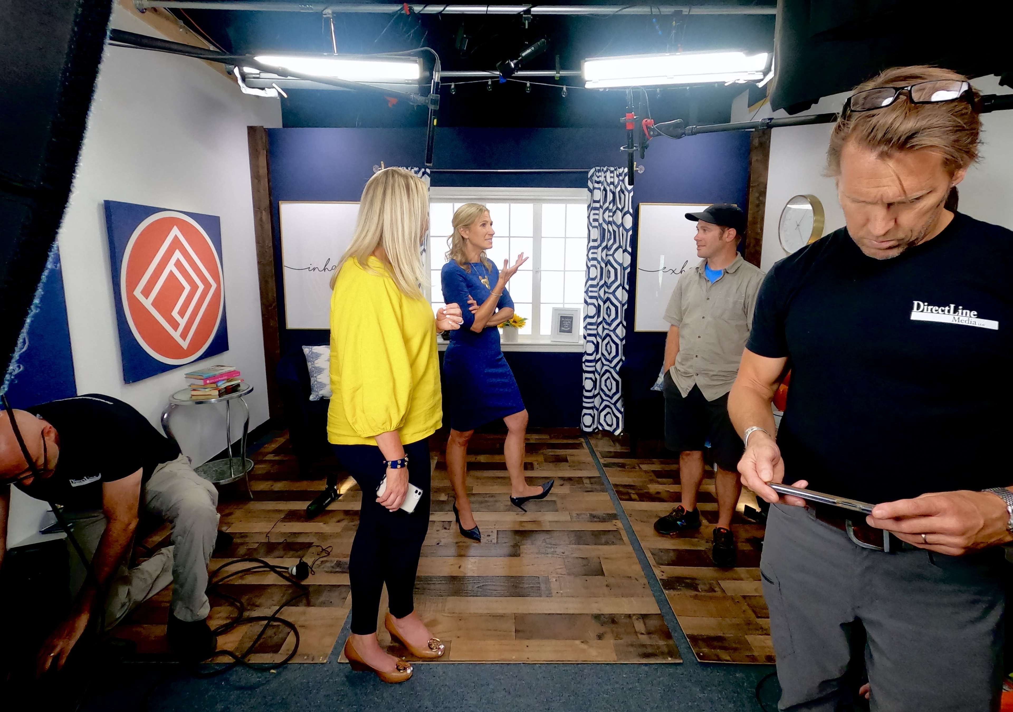 A television production set features a host speaking with a guest while crew members prepare for the shoot.
