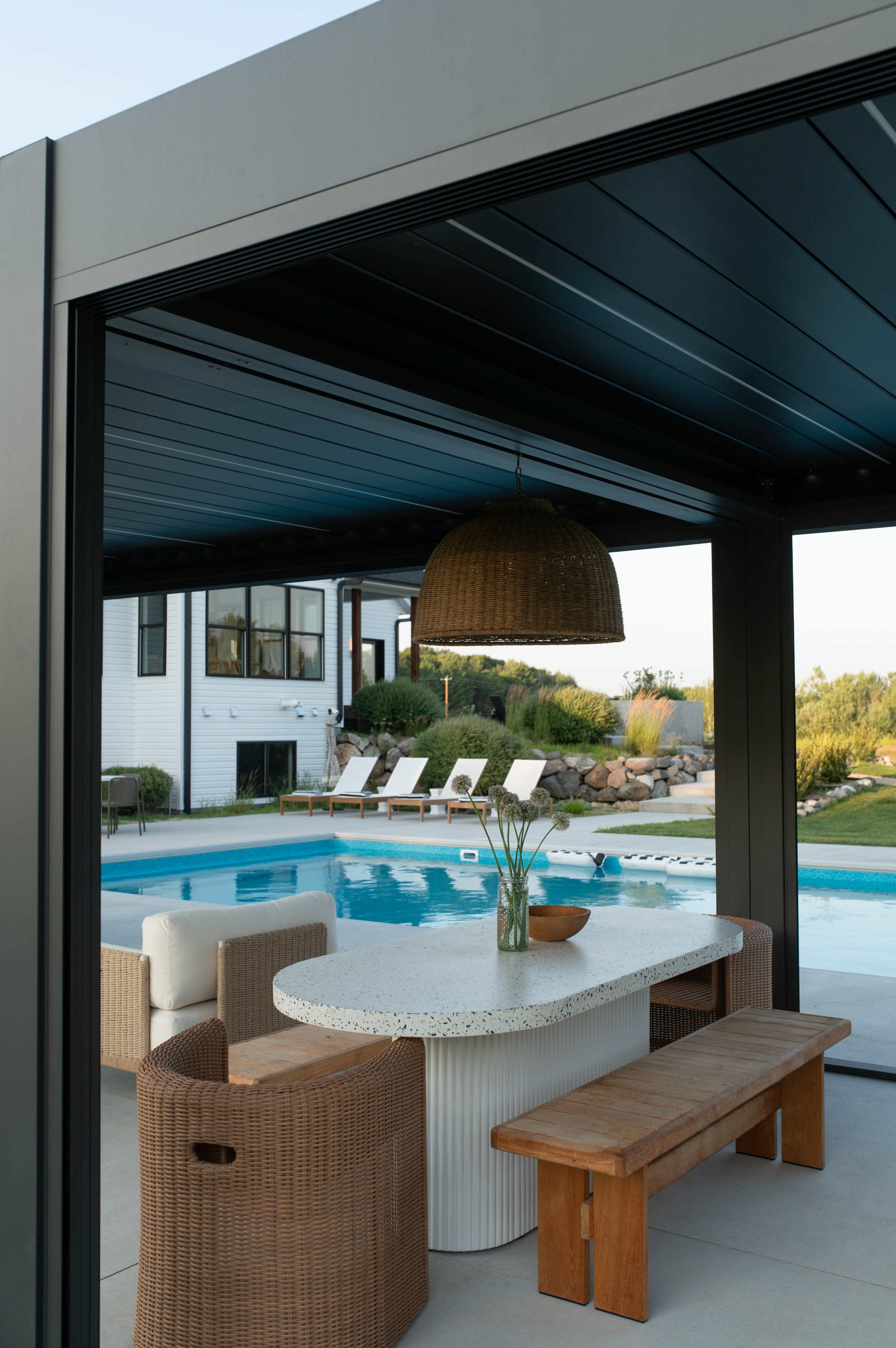 The image shows a patio area with a circular table, a wooden bench, and wicker chairs overlooking a swimming pool surrounded by sun loungers and landscaping.
