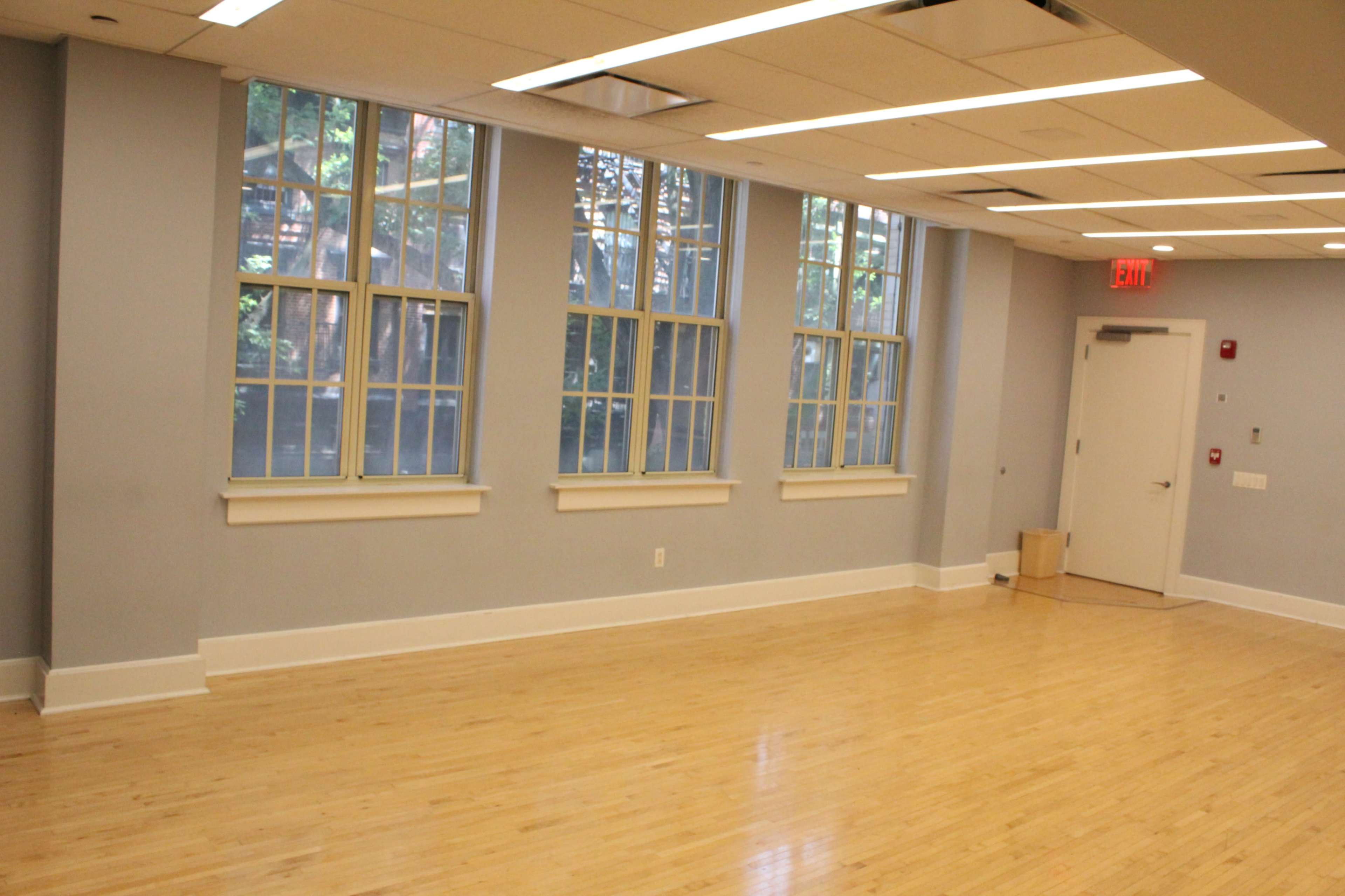 The image shows a vacant room with large windows, wooden flooring, and a door leading outside.