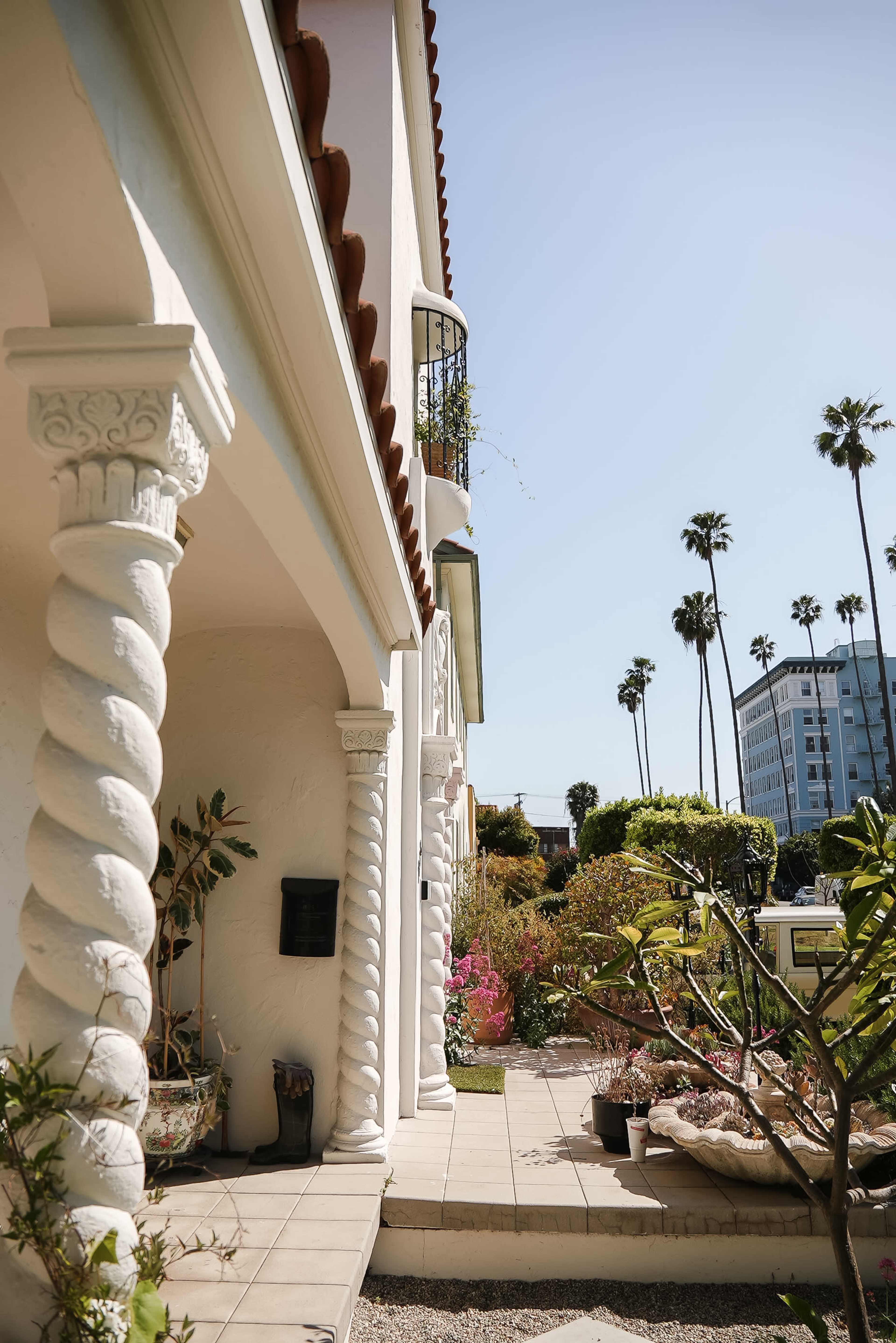 A white building with decorative columns and a garden area, accompanied by palm trees and a multi-story building in the background.