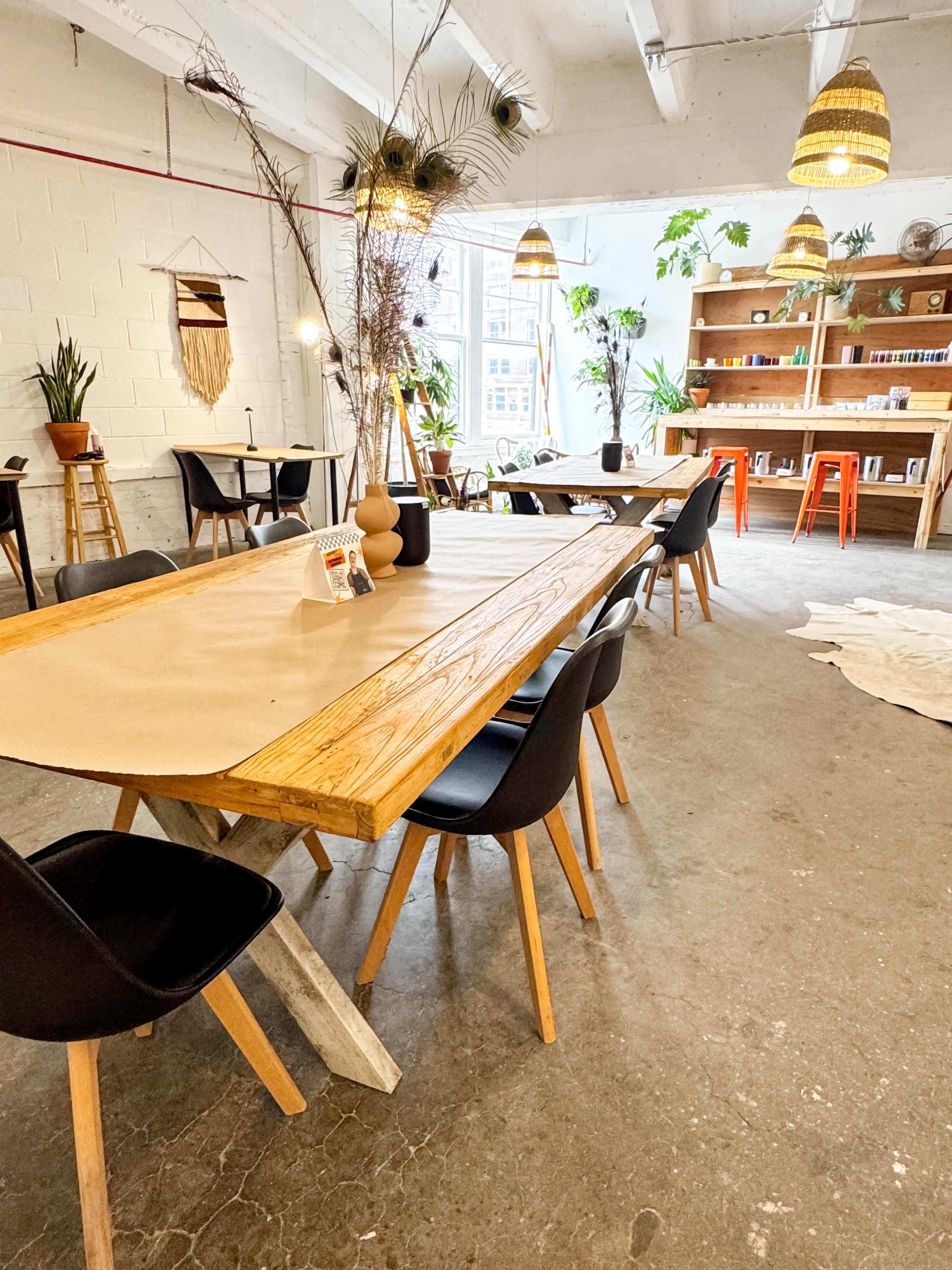 A spacious, well-lit room features a large wooden table surrounded by black chairs, with plants and shelves lining the walls.