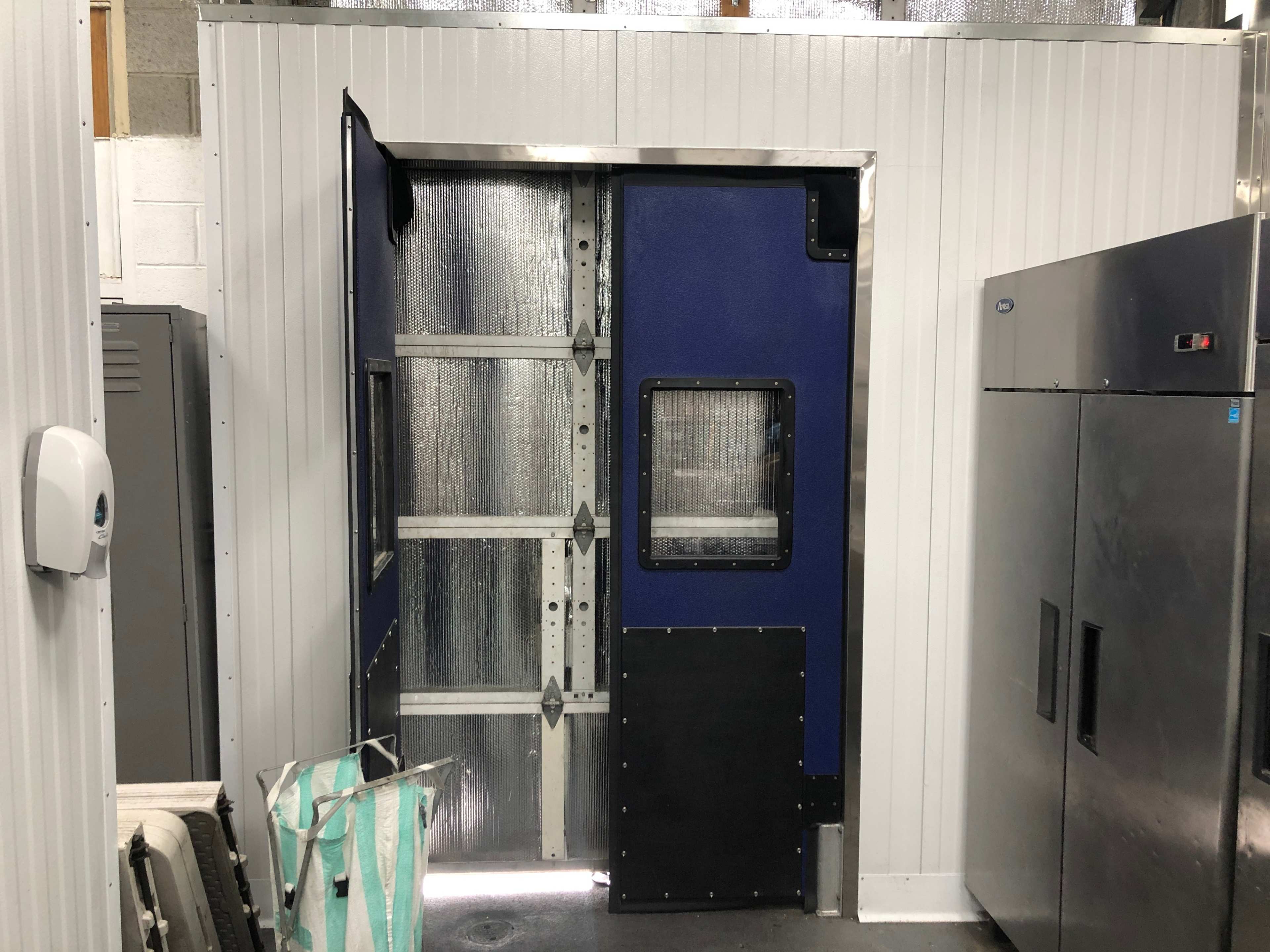 The image shows a blue and silver industrial door set in a metal-paneled wall, adjacent to a stainless steel refrigerator.