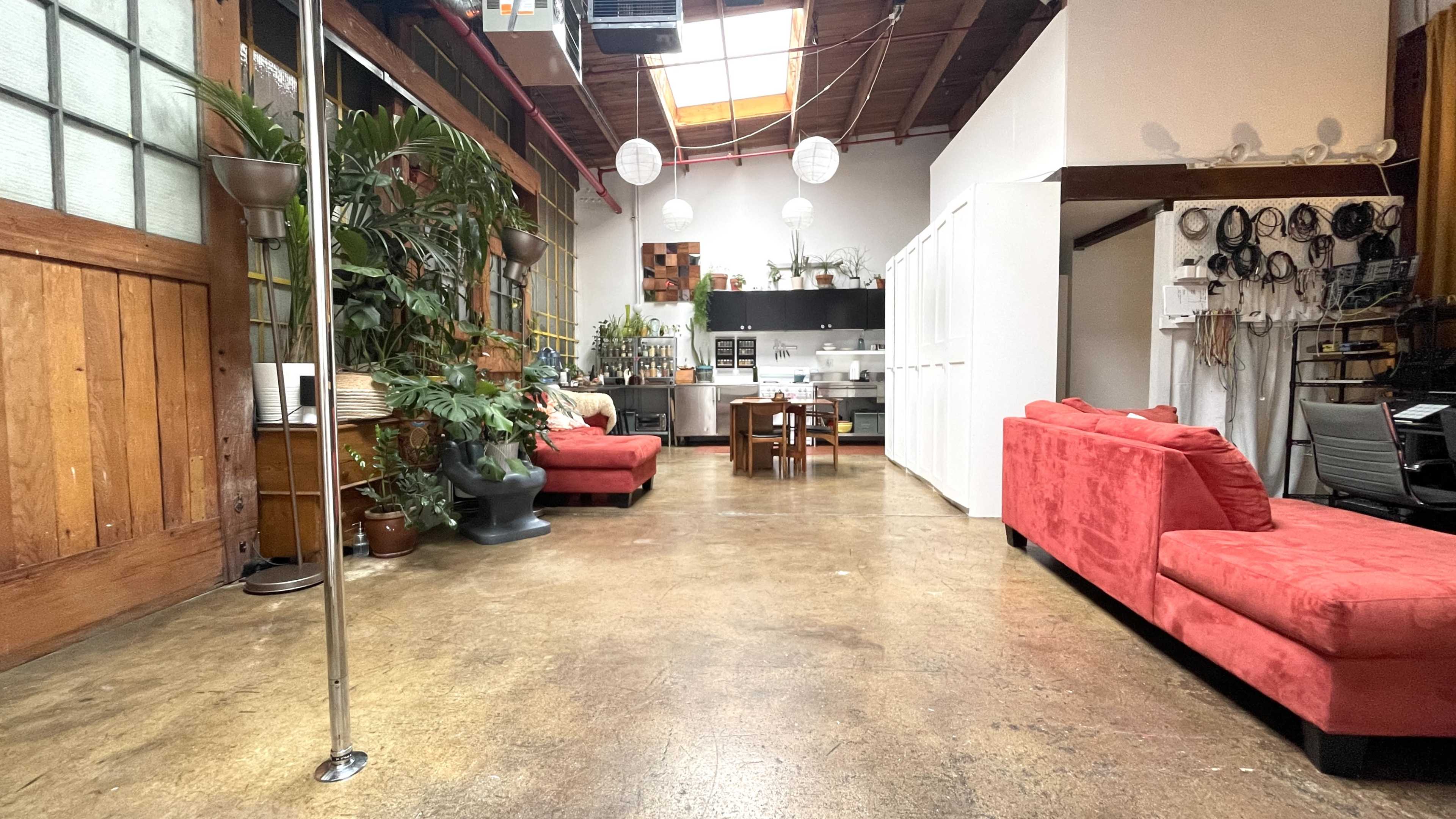The image shows an open, spacious room with wooden beams and a concrete floor, featuring red sofas, plants, and a kitchen area at the back.