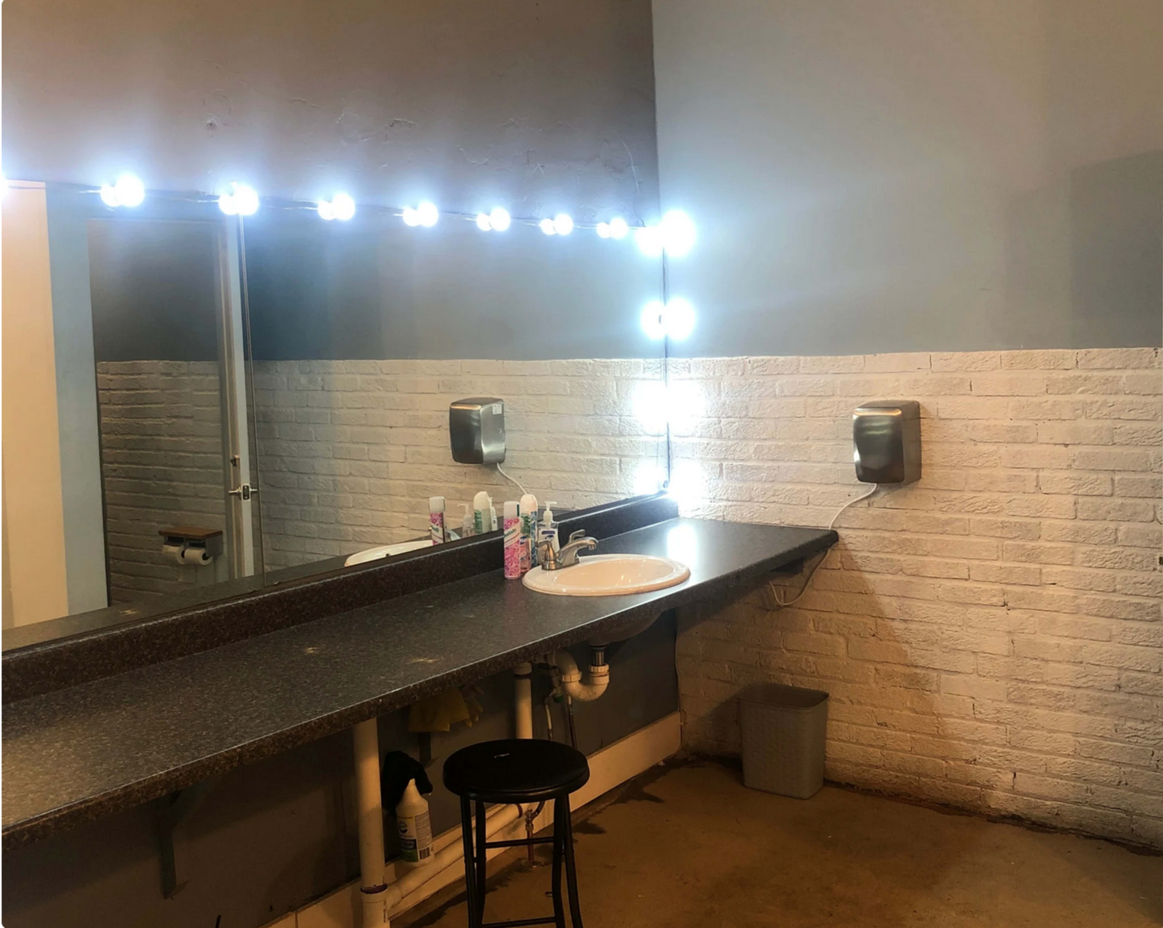 The image displays a bathroom with a long countertop, a circular mirror illuminated by LED lights, and a black stool beside a sink.