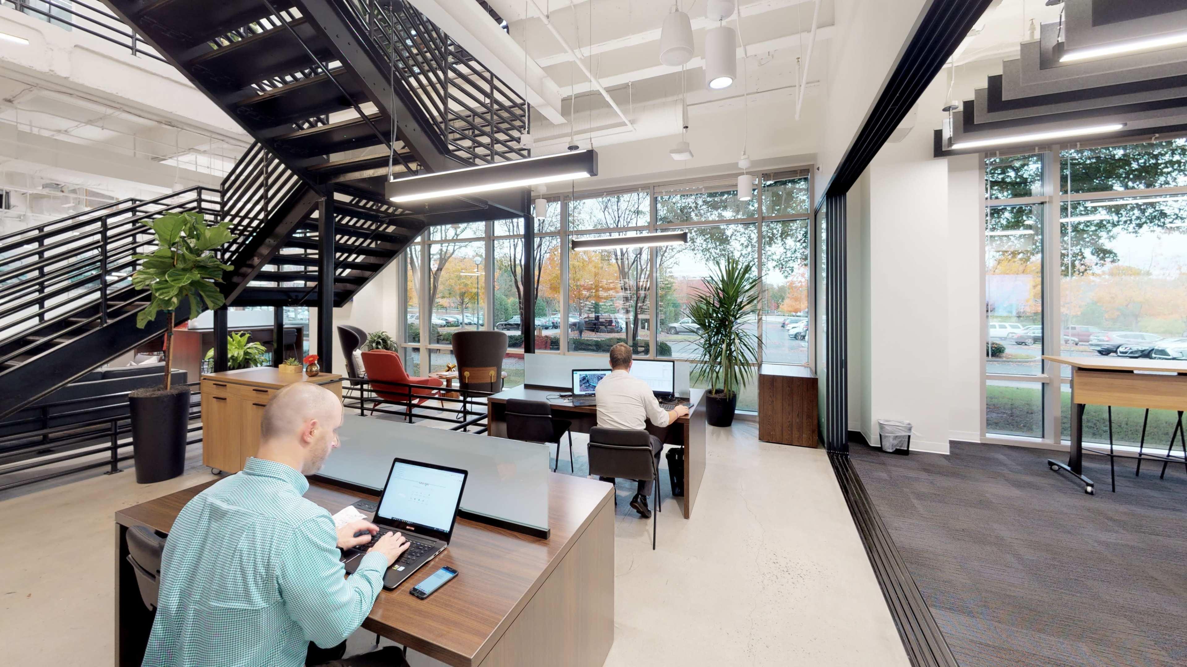 Two individuals are seated at desks in a modern office space, with large windows overlooking a parking lot and a staircase in the background.