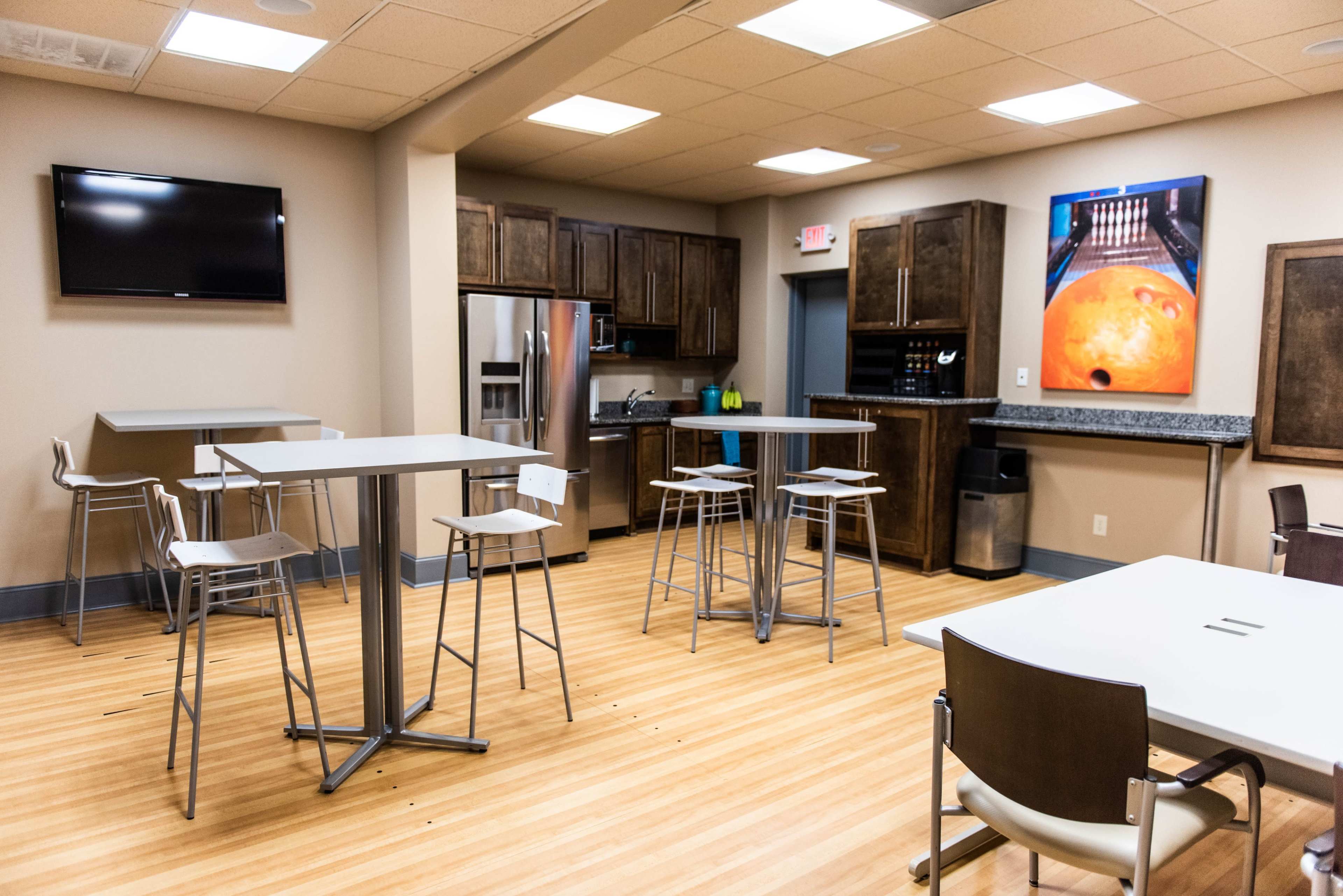 A modern break room featuring several tall tables and stools, a kitchenette with dark wood cabinets, and a television mounted on the wall.