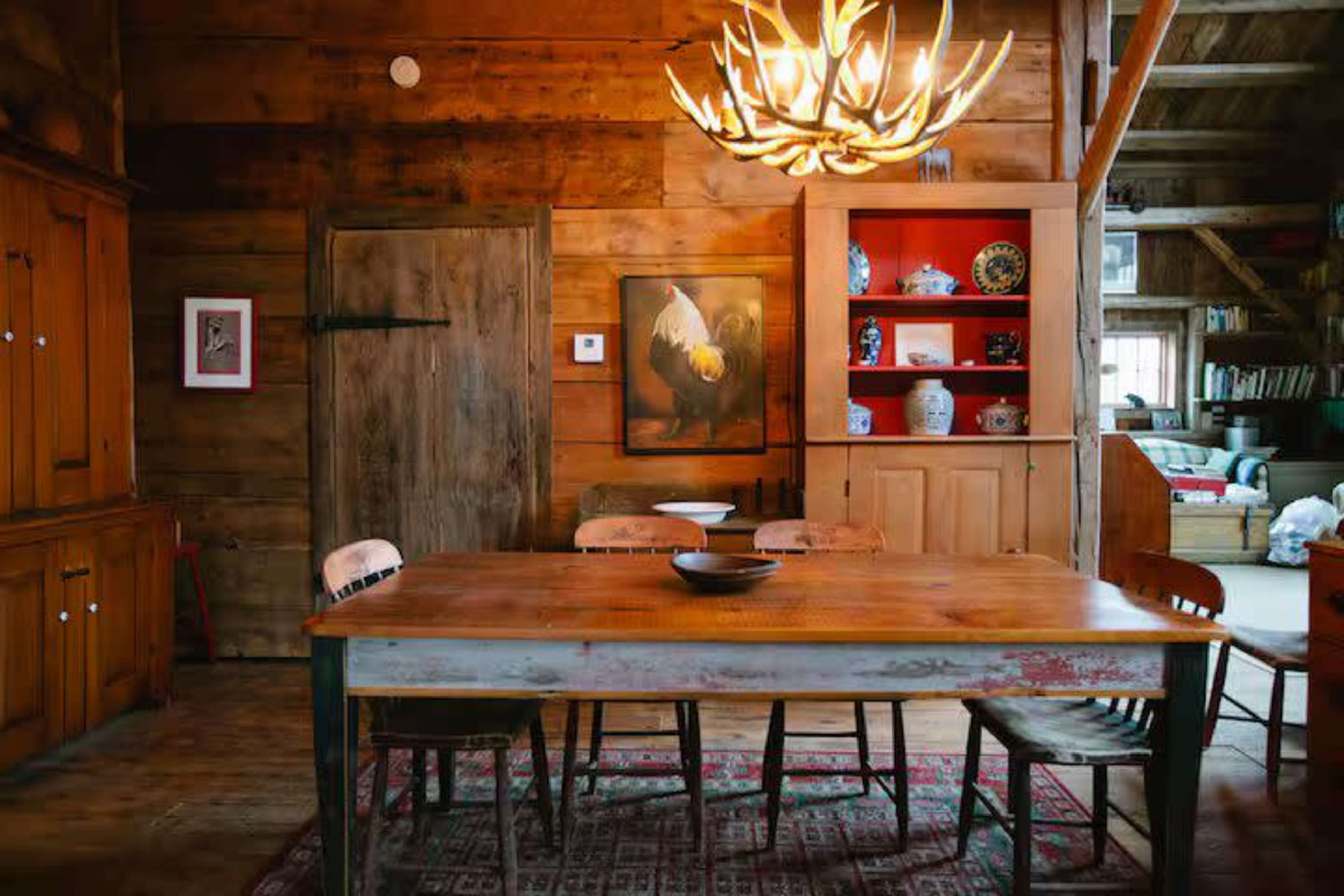 The image shows a rustic dining room featuring a wooden table surrounded by chairs, a large antler-shaped chandelier, and wooden walls with a variety of decorative items on shelves.
