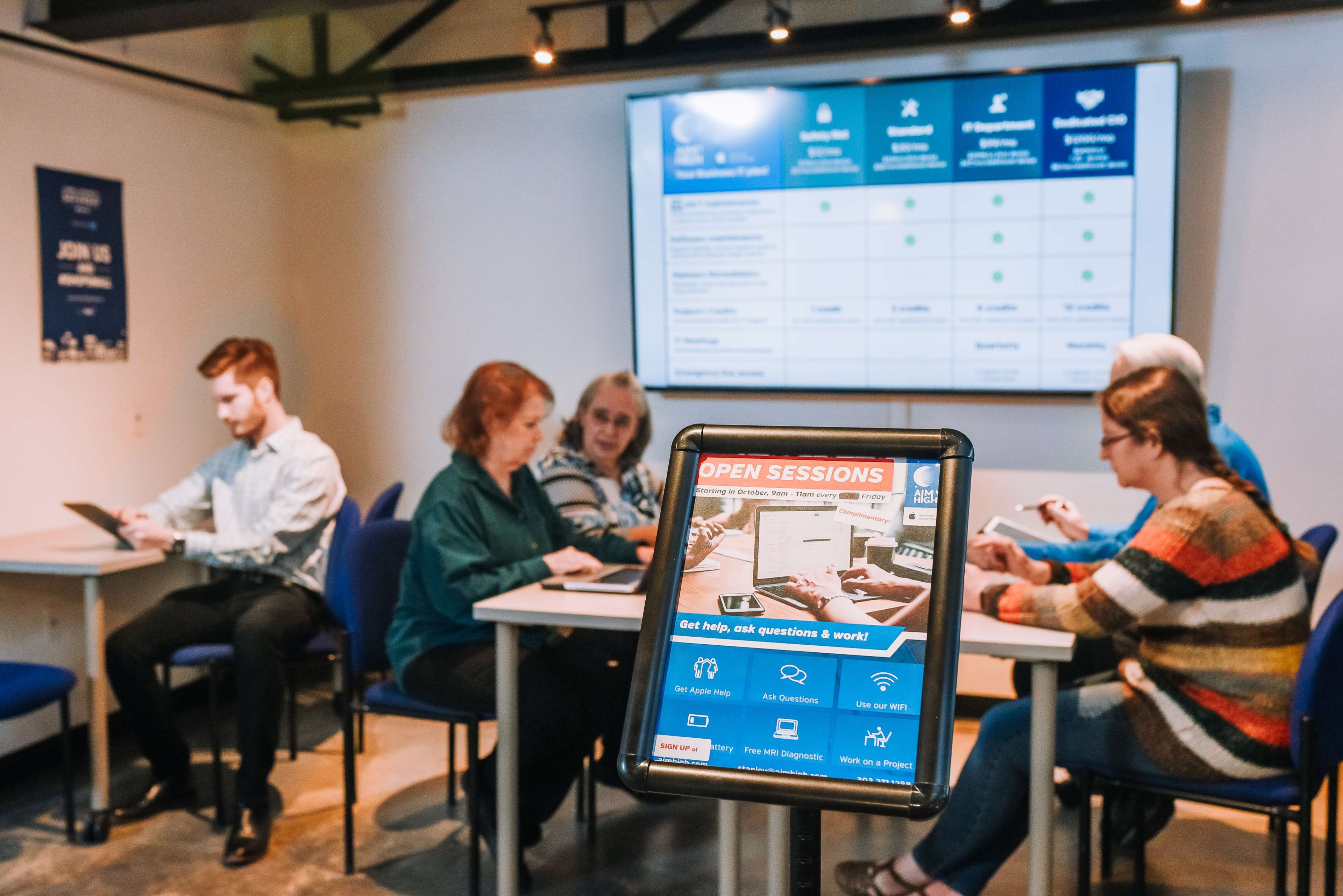 The image shows a group of people engaged in a discussion at a table in a room with a large display board behind them, while a digital sign promotes open sessions for asking questions.