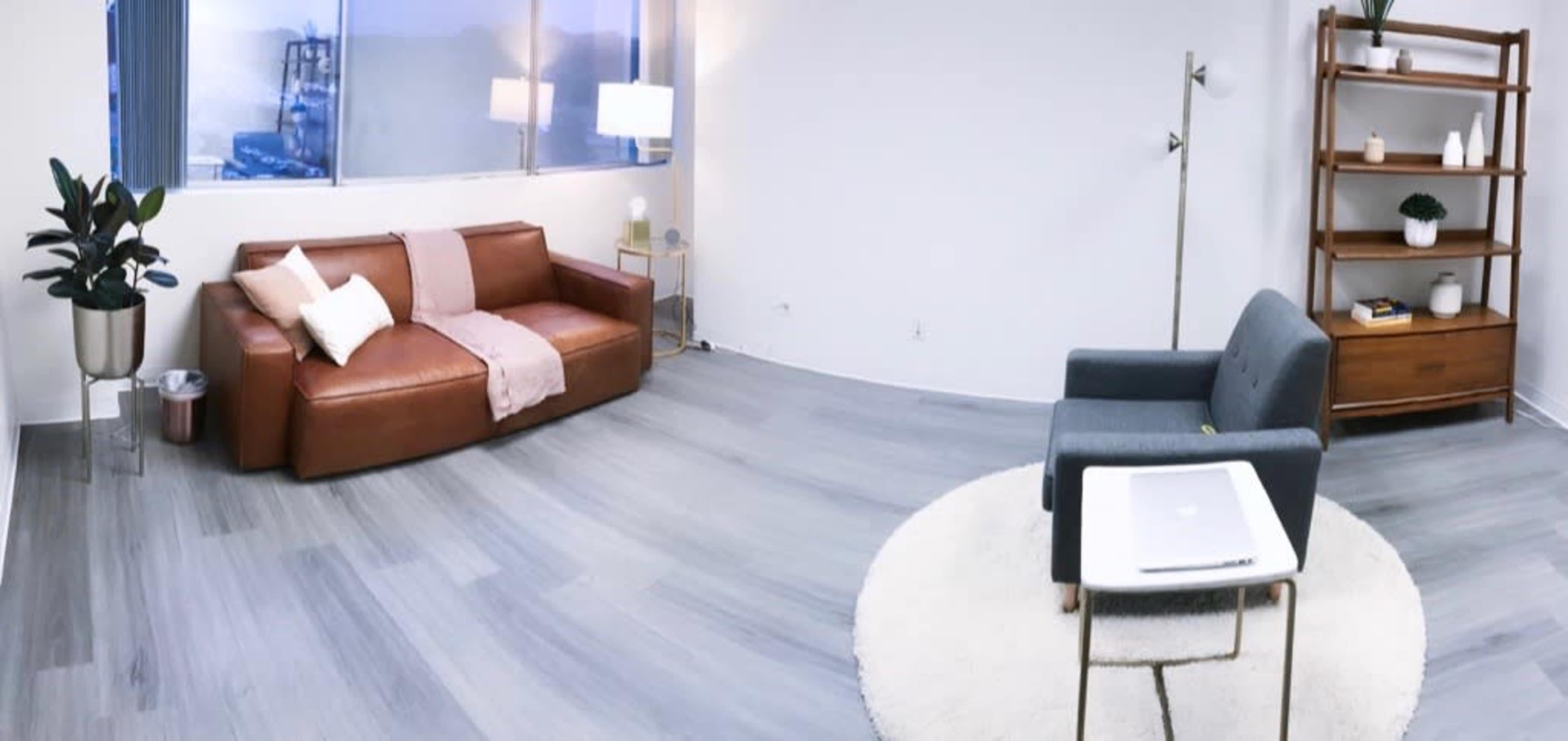 A modern, minimalist living space featuring a brown leather sofa, a gray armchair, a round white rug, a side table, and a shelving unit against a wall with windows allowing natural light.
