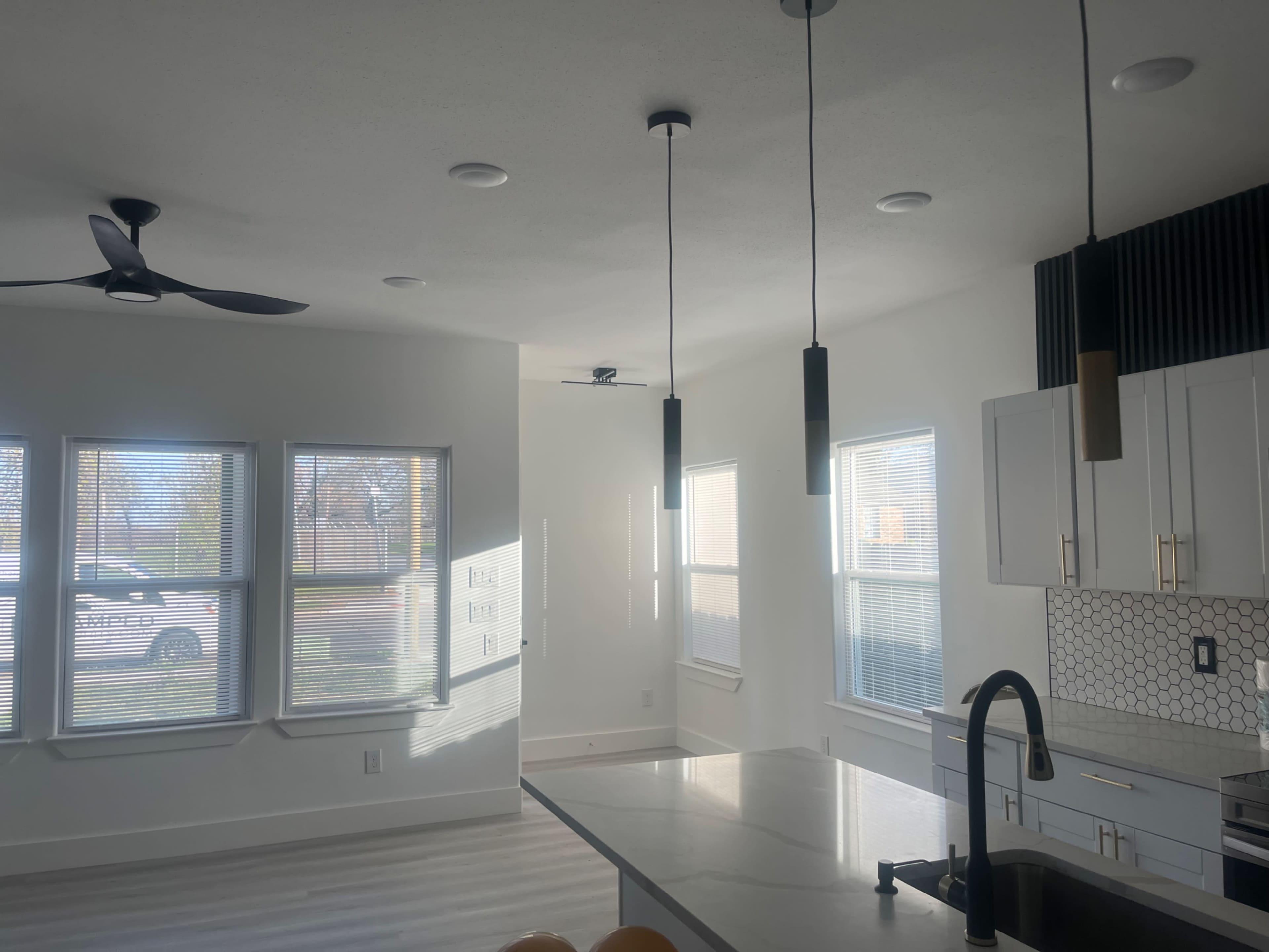 The image shows a modern kitchen and living area with large windows allowing natural light, featuring a countertop, pendant lighting, and minimalist decor.