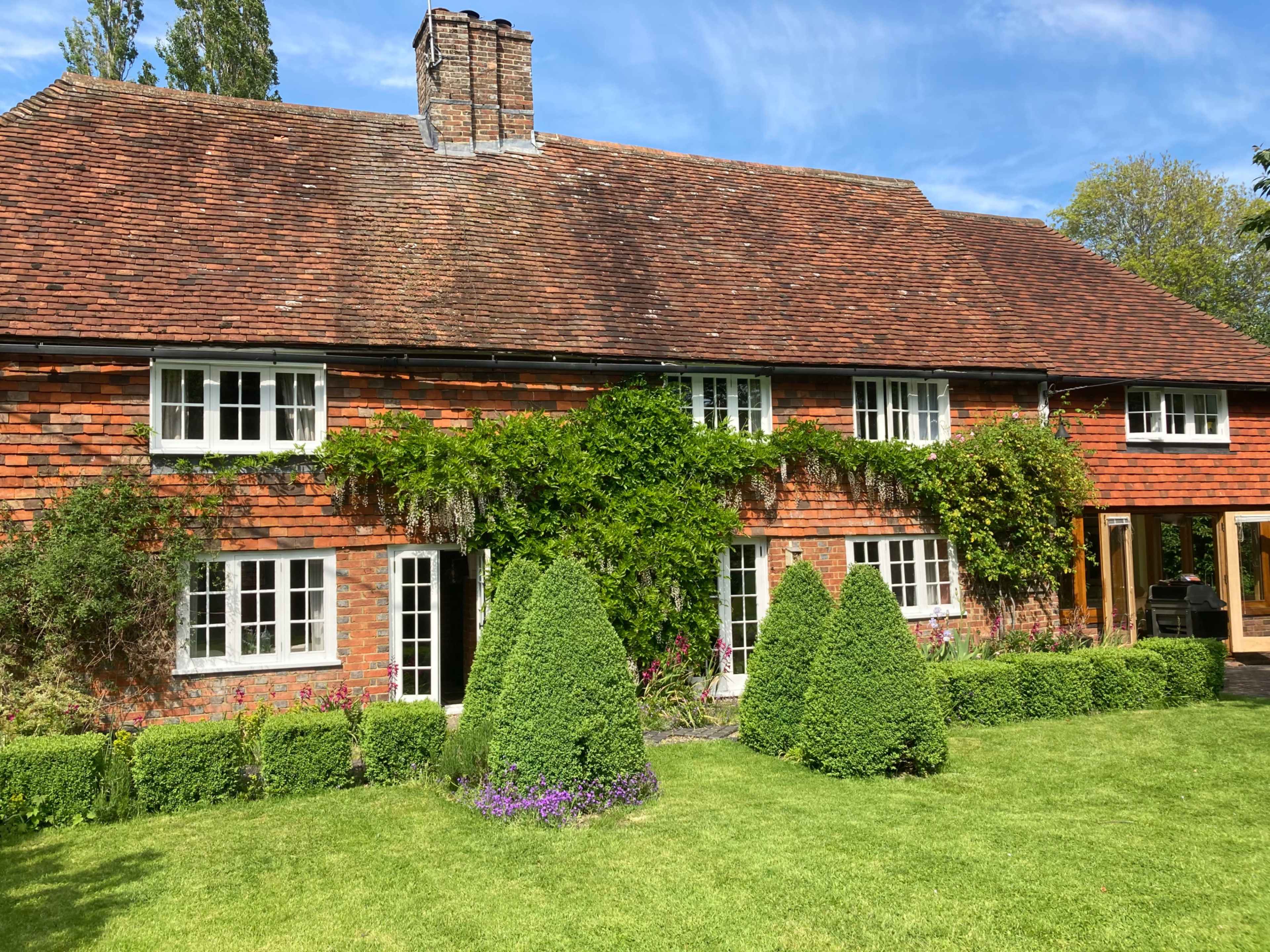 A brick house with a sloped roof, surrounded by well-maintained green shrubs and a lawn, with trees in the background under a blue sky.