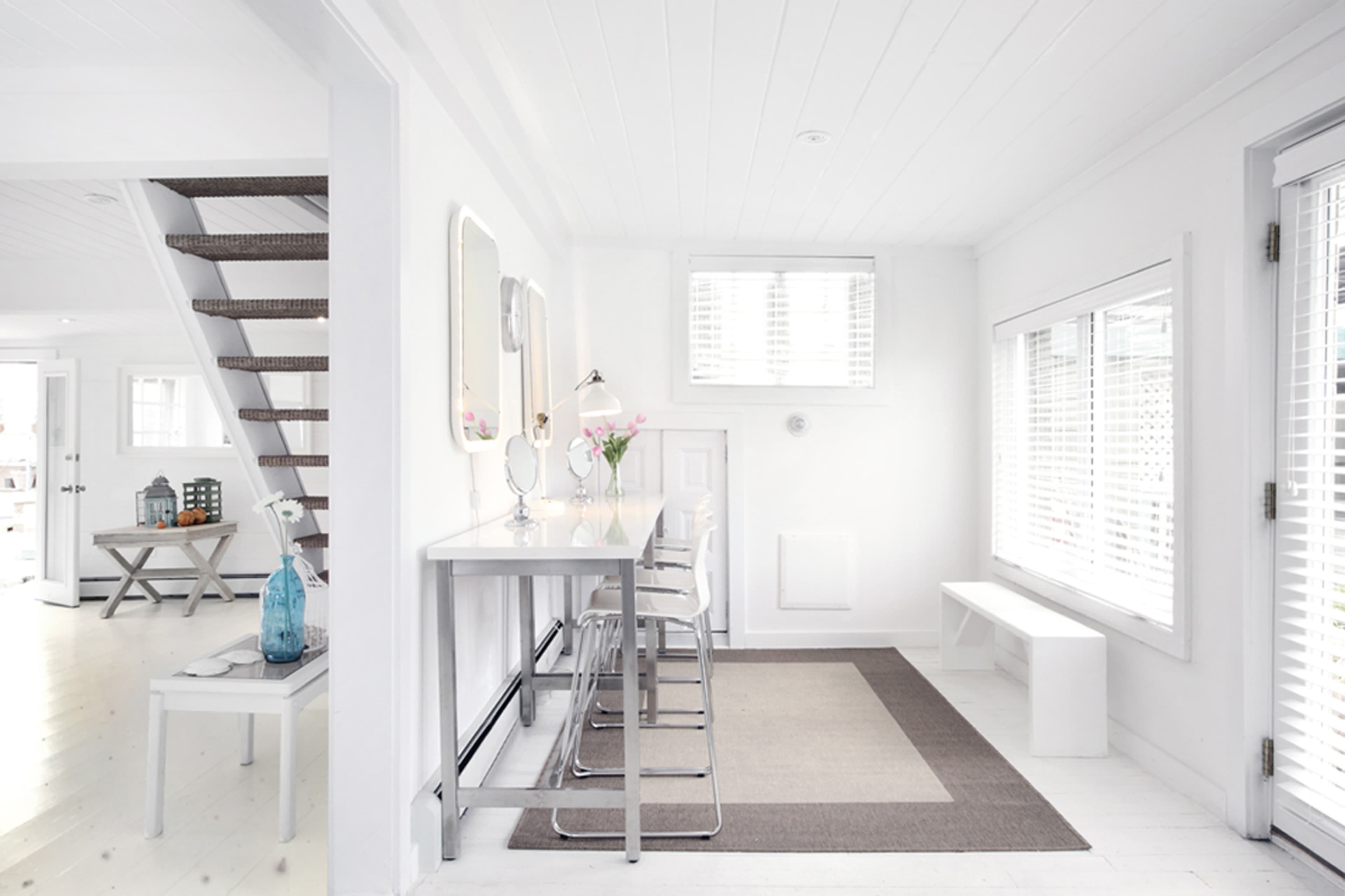 A bright, minimalist interior featuring a dining area with a table and chairs, a staircase, and large windows with blinds.