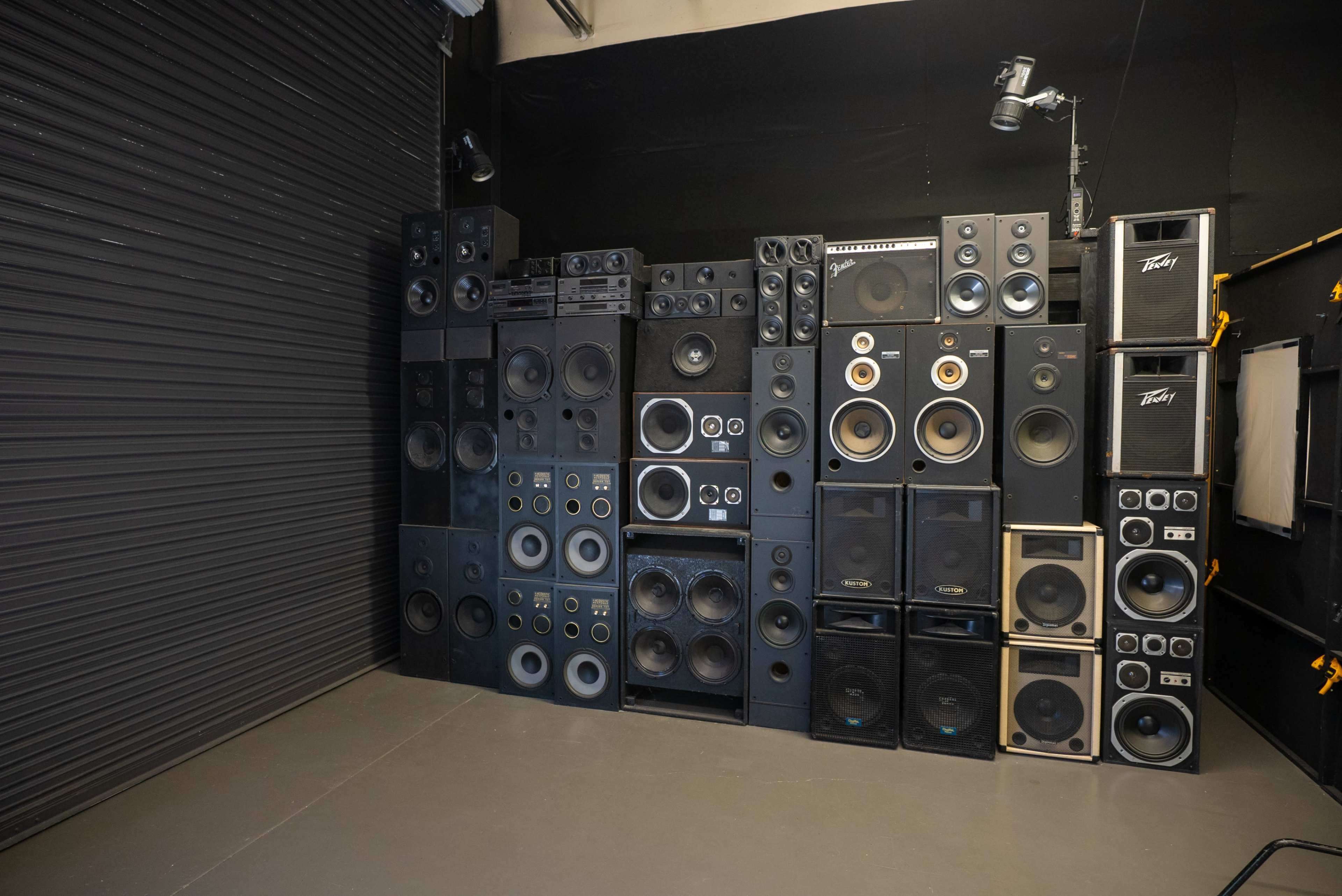 A wall of large audio speakers is stacked against a black backdrop in a studio setting.