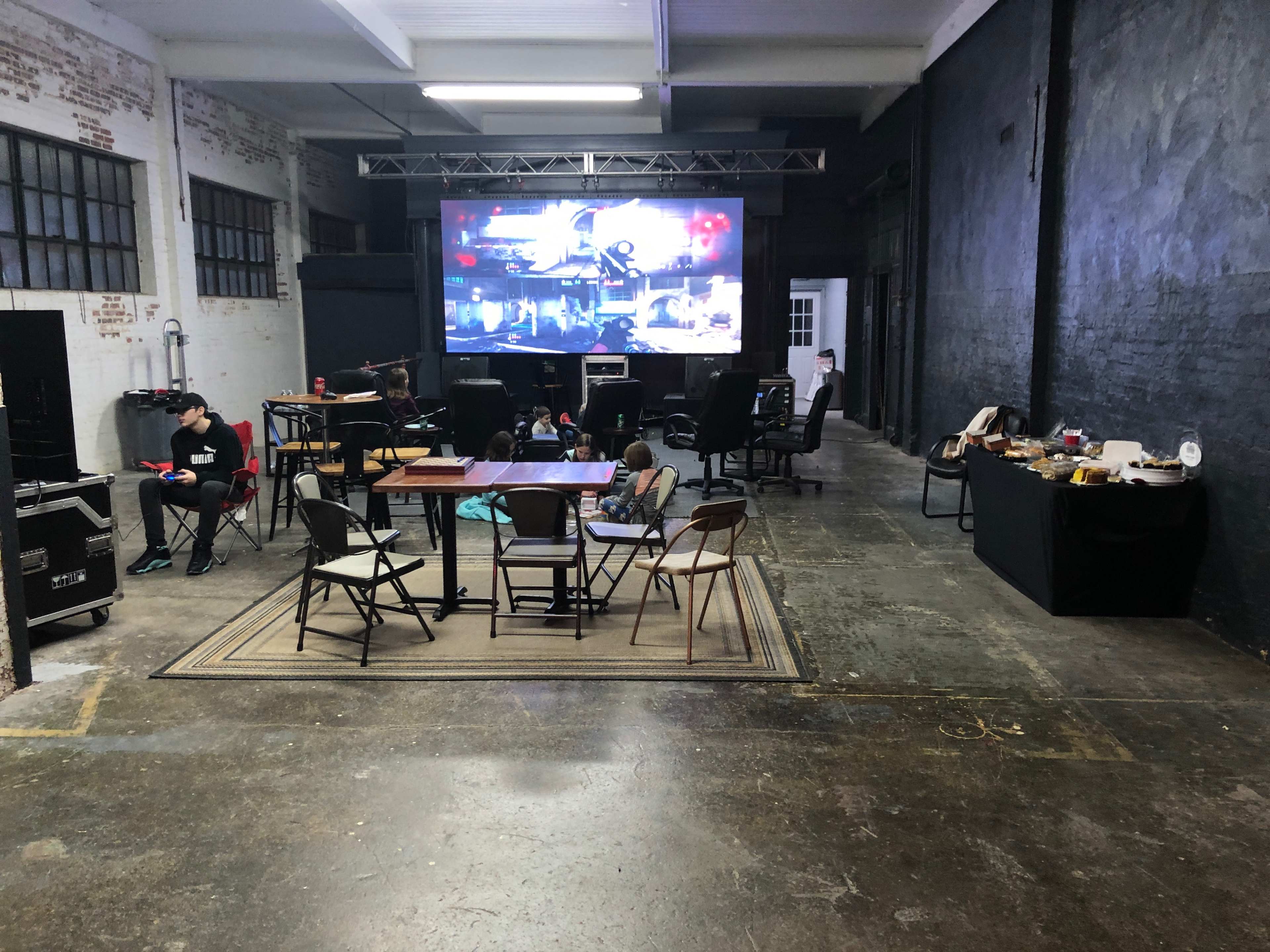 A large screen displays images in an industrial-style room with chairs and tables arranged around a carpet.