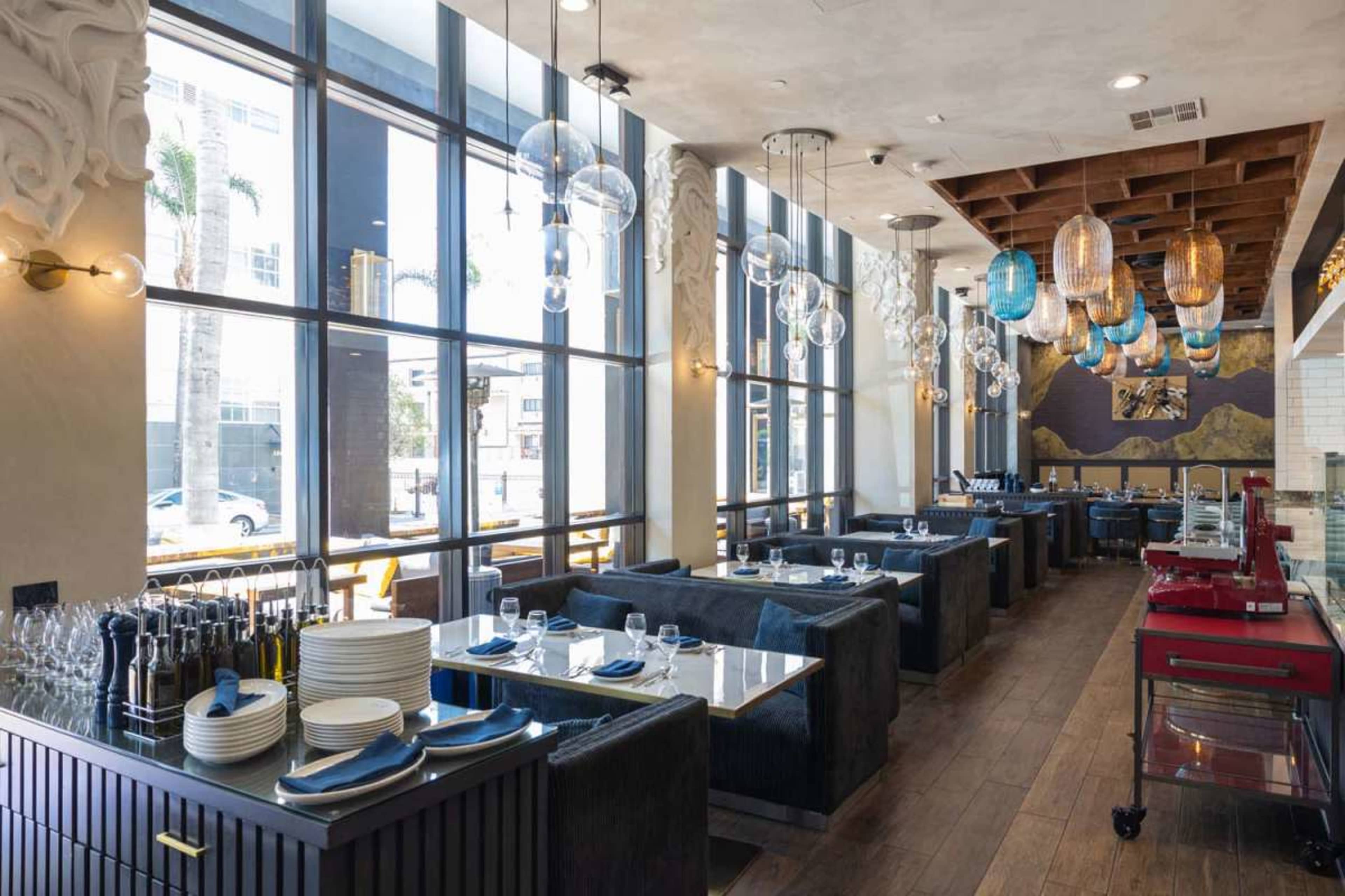 The image shows the interior of a restaurant featuring large windows, wooden beams on the ceiling, and a mix of booth seating and tables, with various light fixtures hanging overhead.