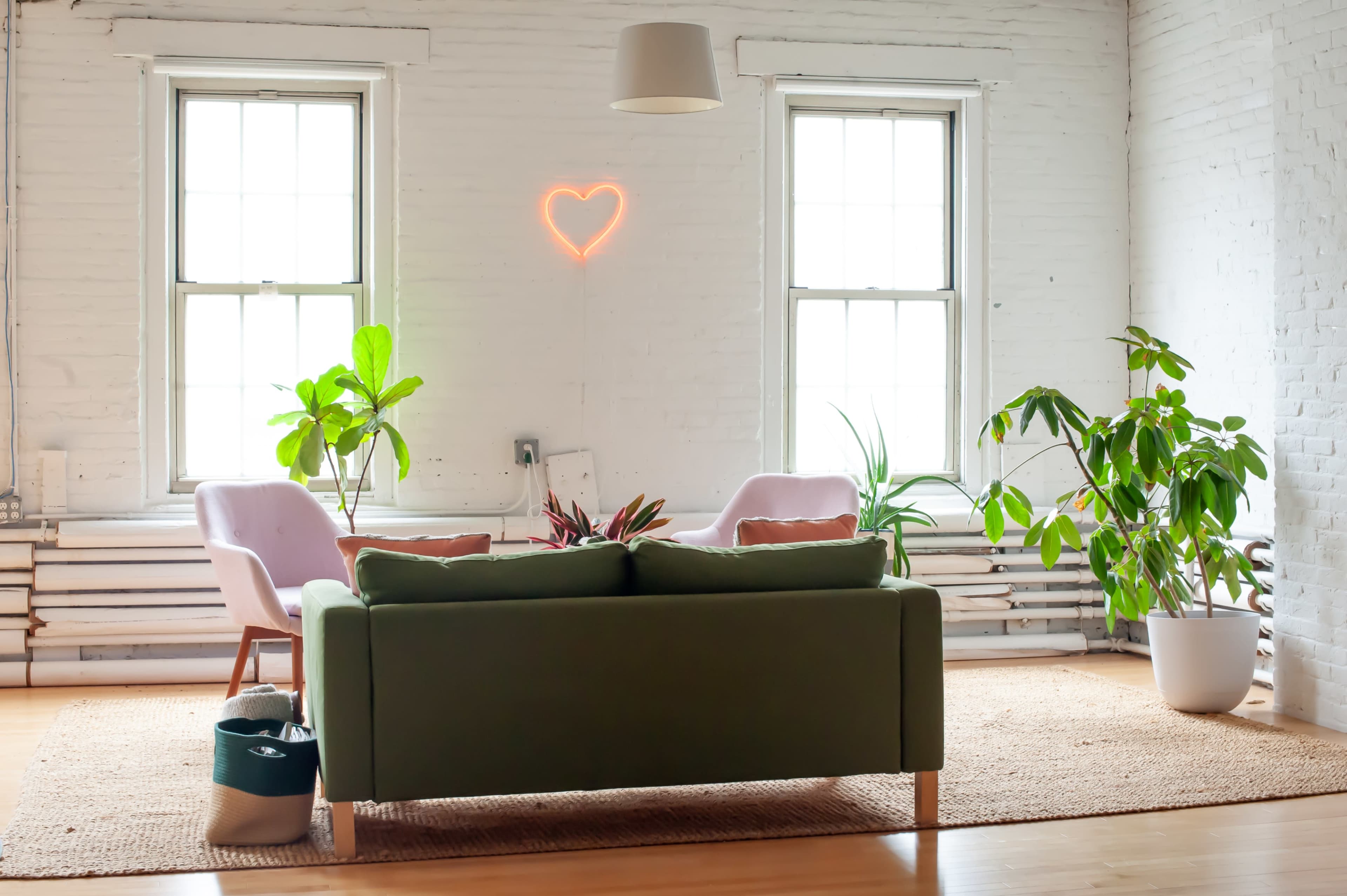 The image shows a minimalist living room with a green sofa, two armchairs, houseplants, and a heart-shaped neon light on the wall.