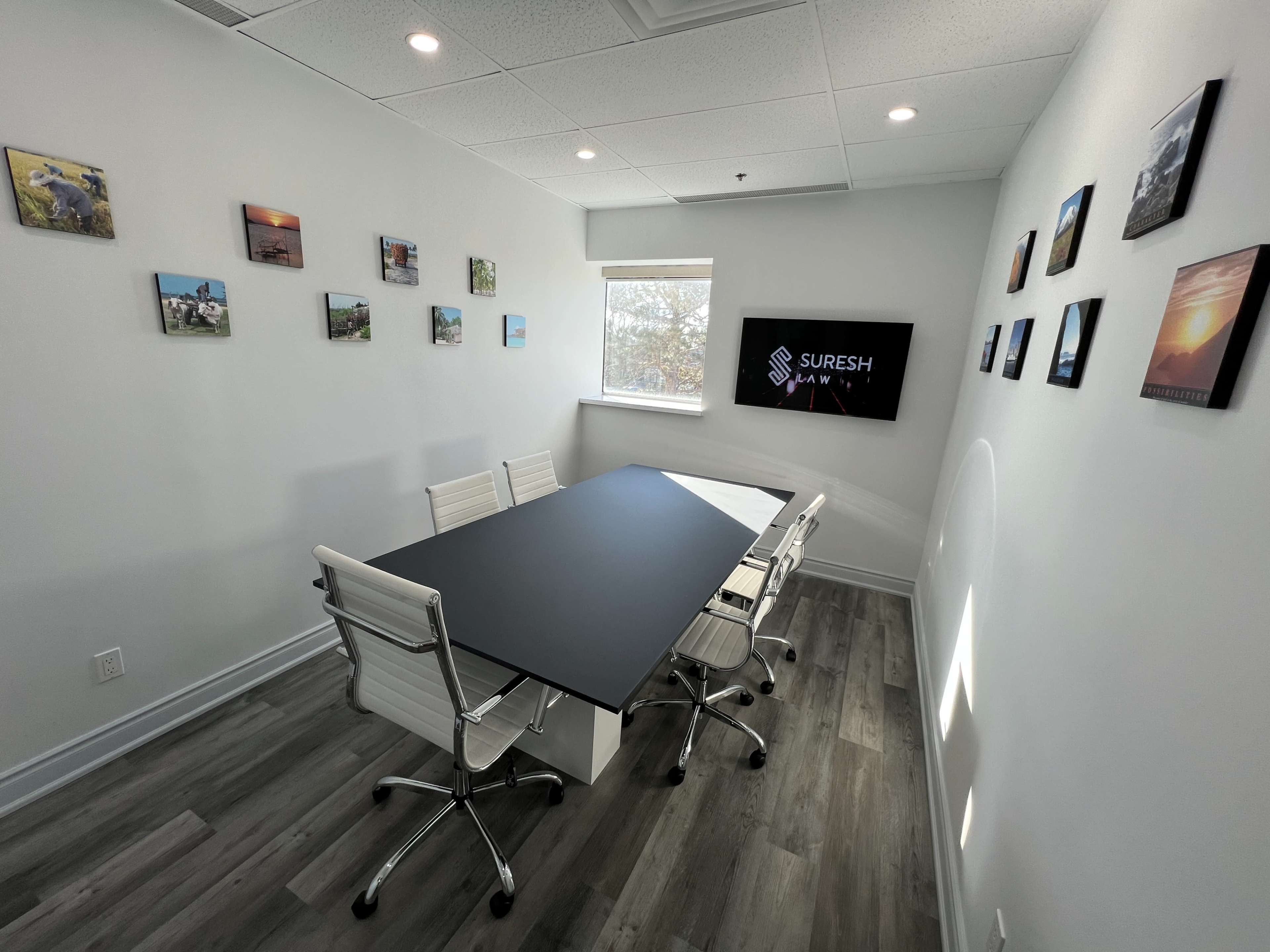 A modern conference room features a rectangular table surrounded by white chairs, with artwork on the walls and a display screen showing the name "Suresh."