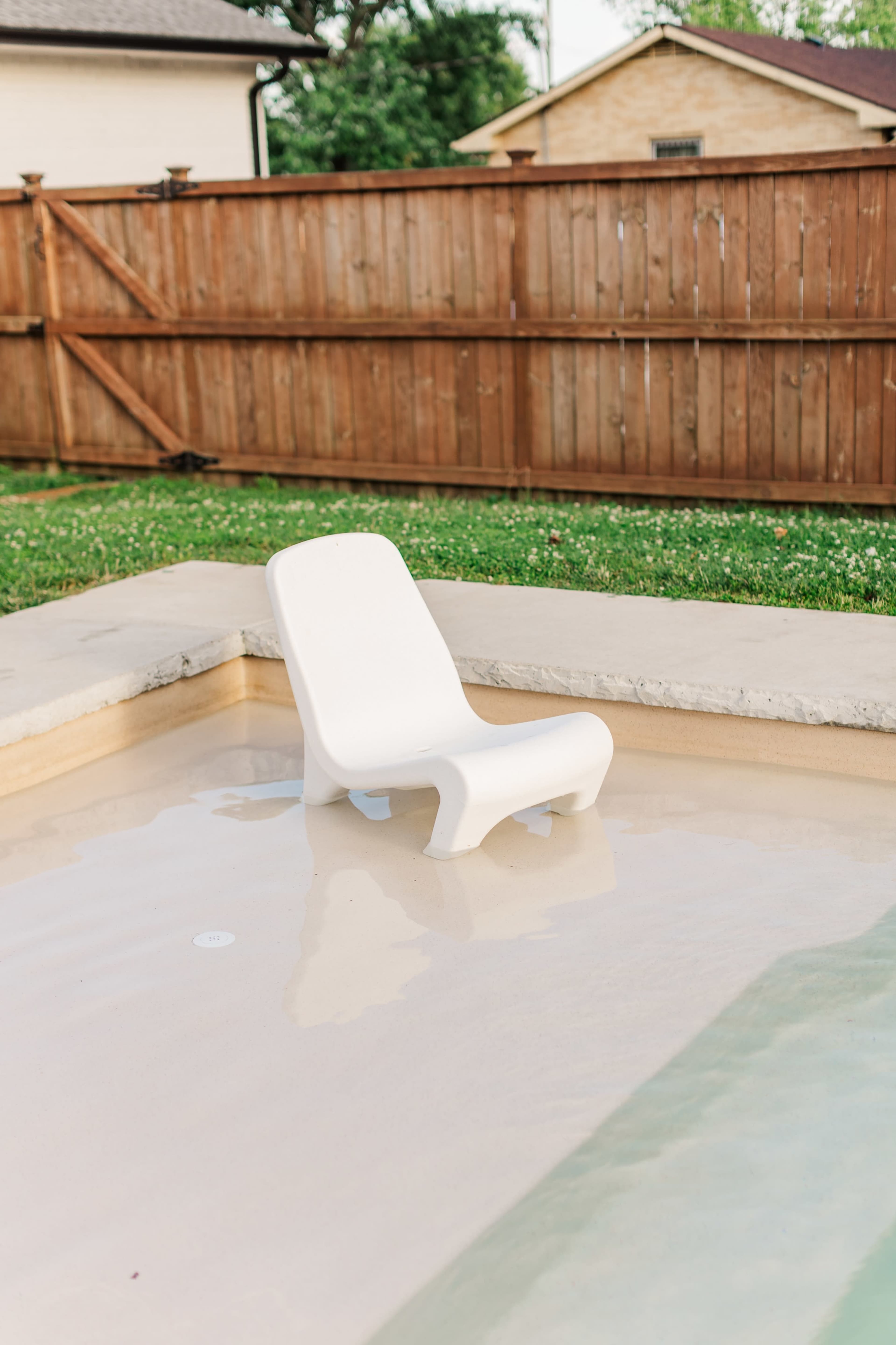 A white plastic chair is sitting in shallow water at the edge of a swimming pool, surrounded by a grassy area and a wooden fence.