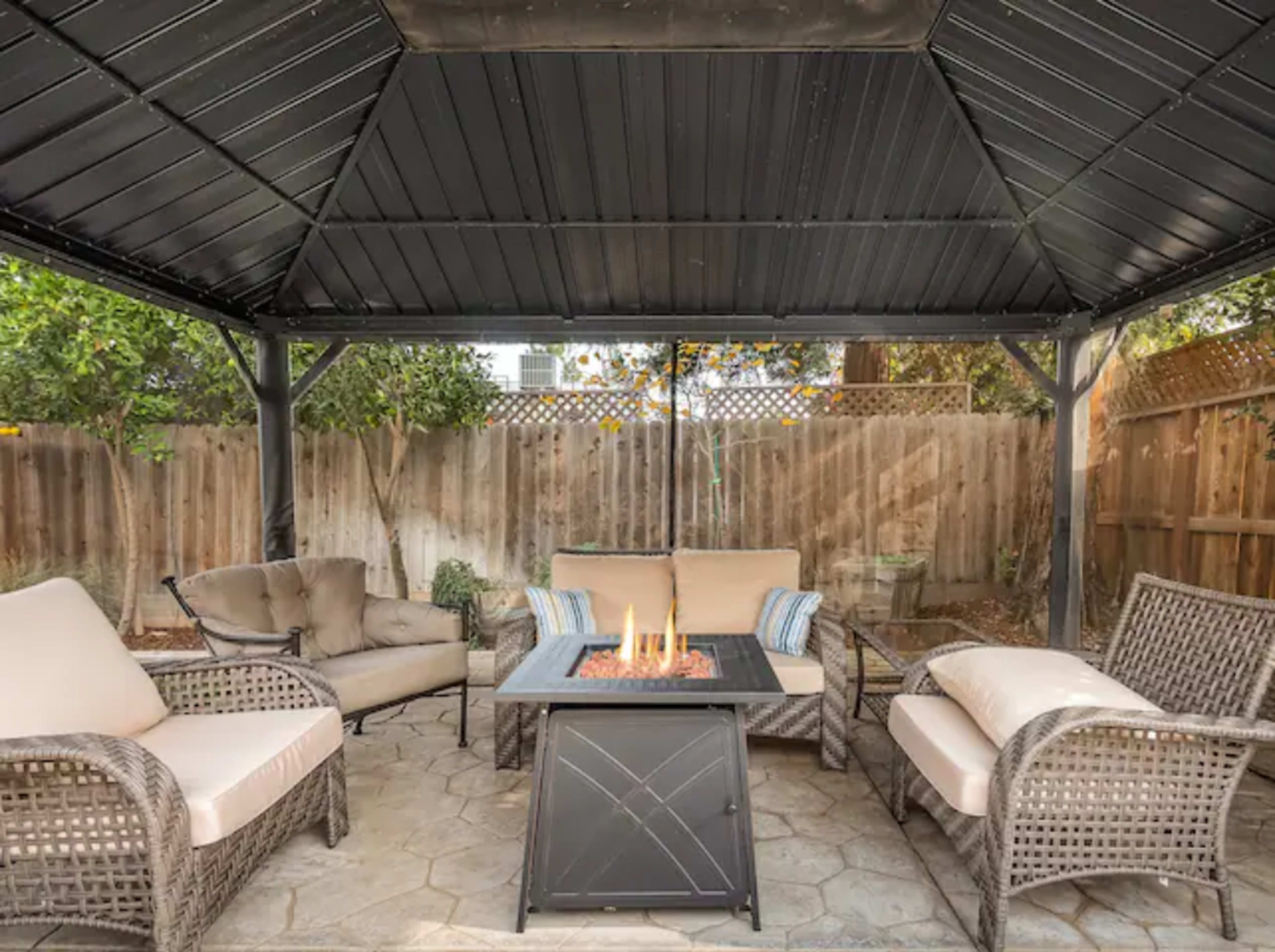 A covered outdoor seating area features a fire pit surrounded by several chairs and a couch on a stone patio.