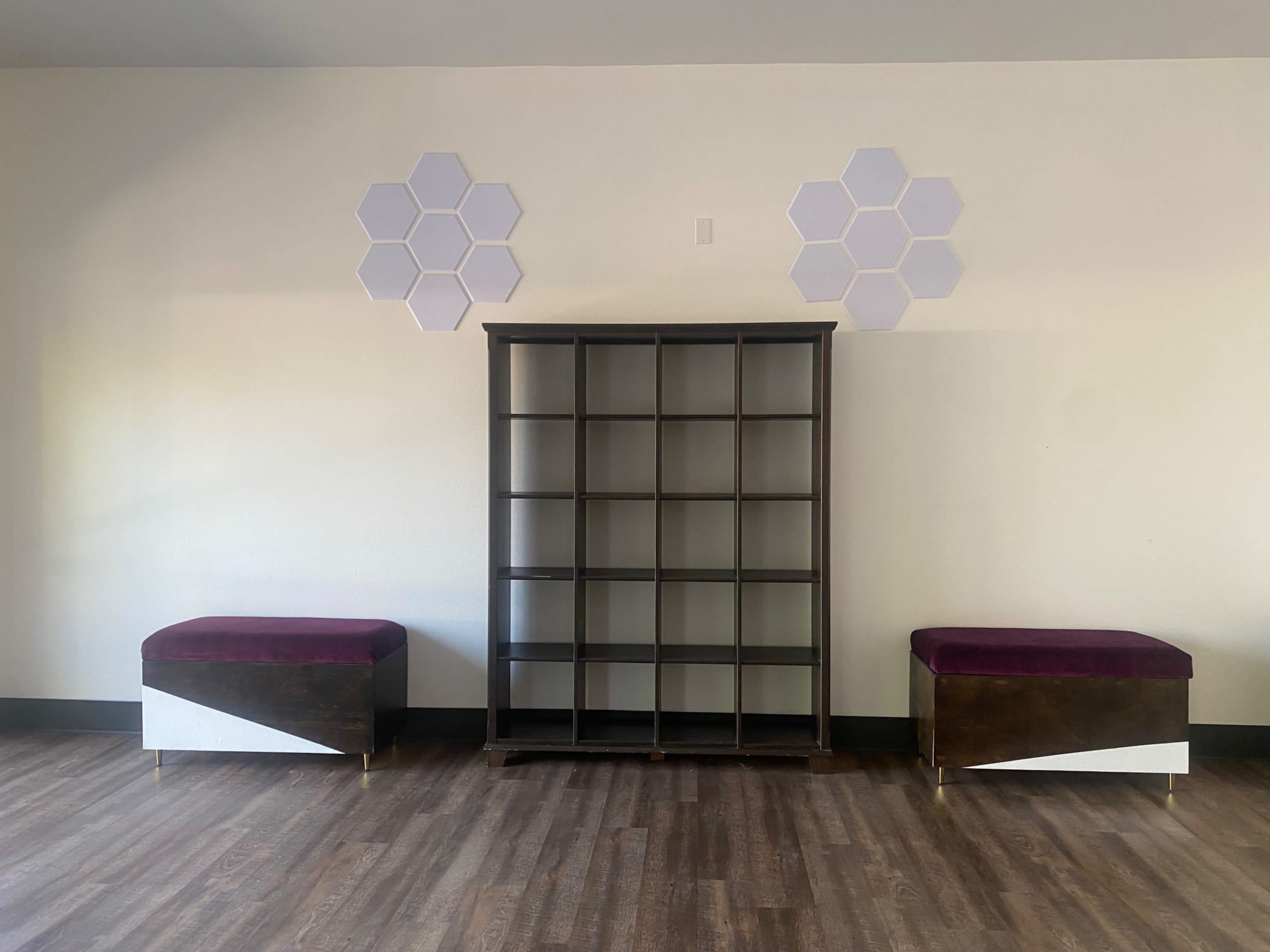 The image shows a wooden bookshelf in the center flanked by two benches with purple upholstery, all set against a plain wall featuring hexagonal wall decorations.