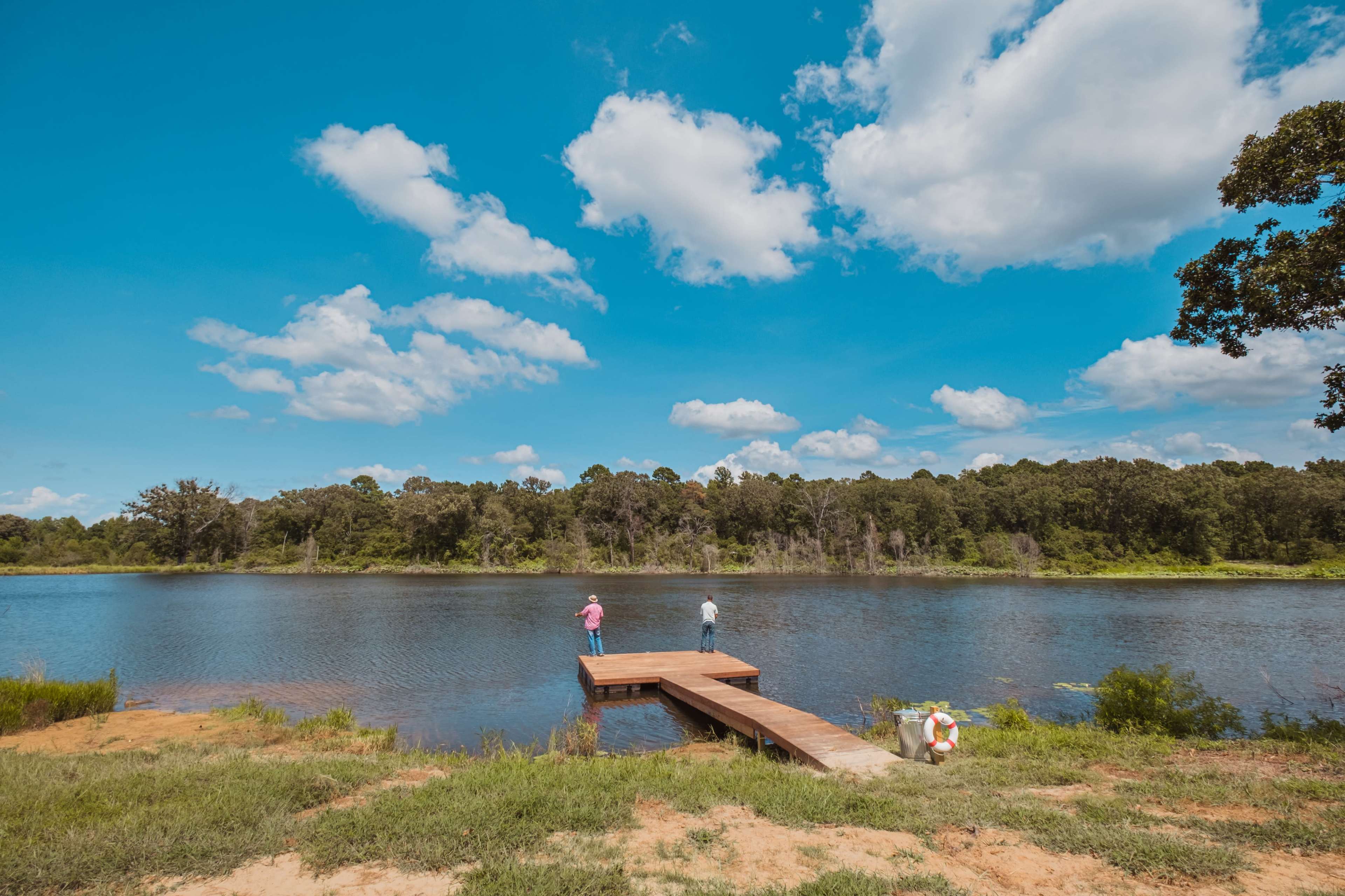Three people stand on a wooden dock by a calm lake under a blue sky filled with clouds.