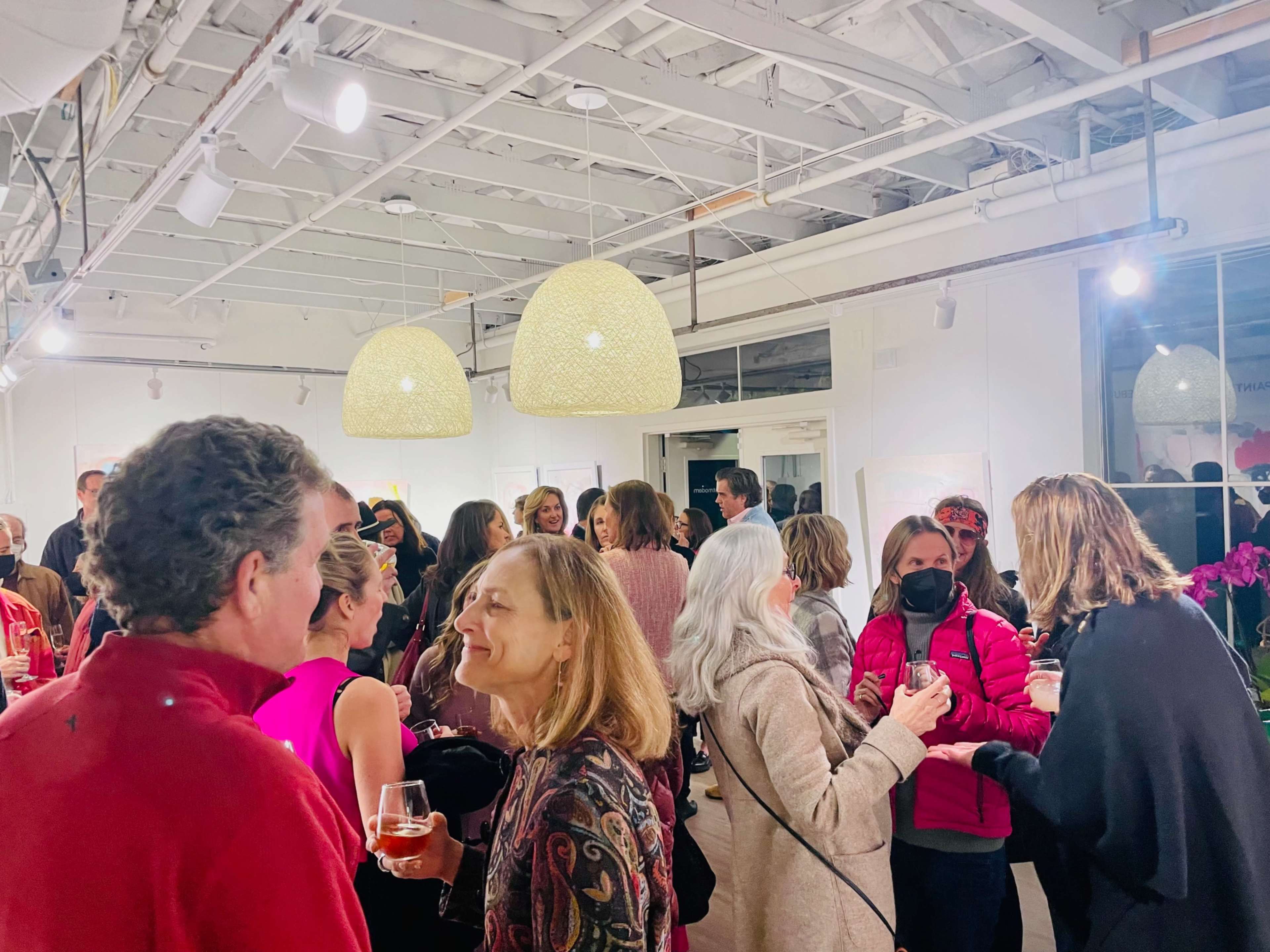 A lively crowd of people mingles and converses at an art gallery event, with pendant lights hanging from the ceiling and decorative plants in the background.