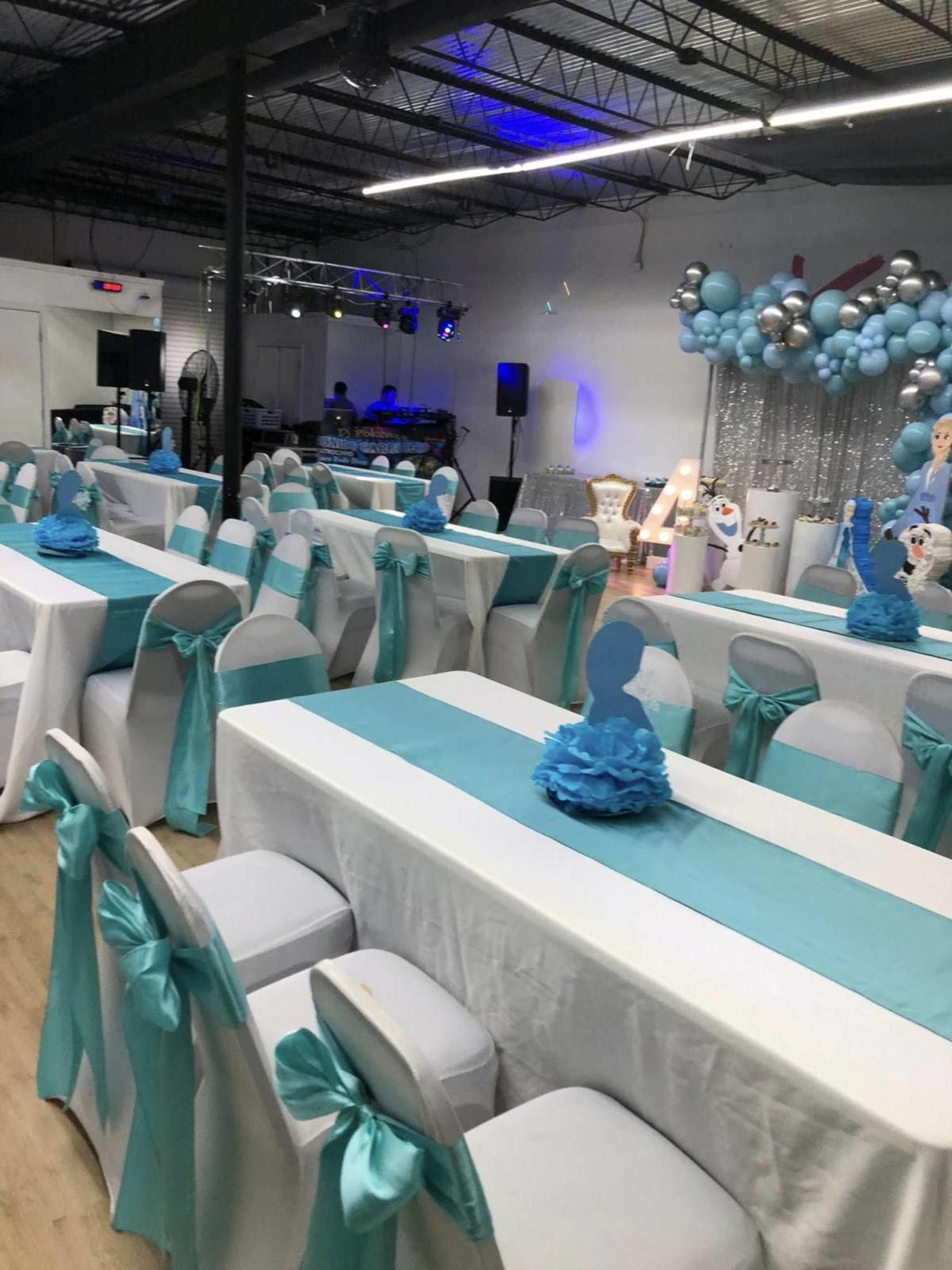 The image shows a decorated event space with rows of tables covered in white linens and turquoise accents, featuring a festive backdrop with balloons and themed decorations.