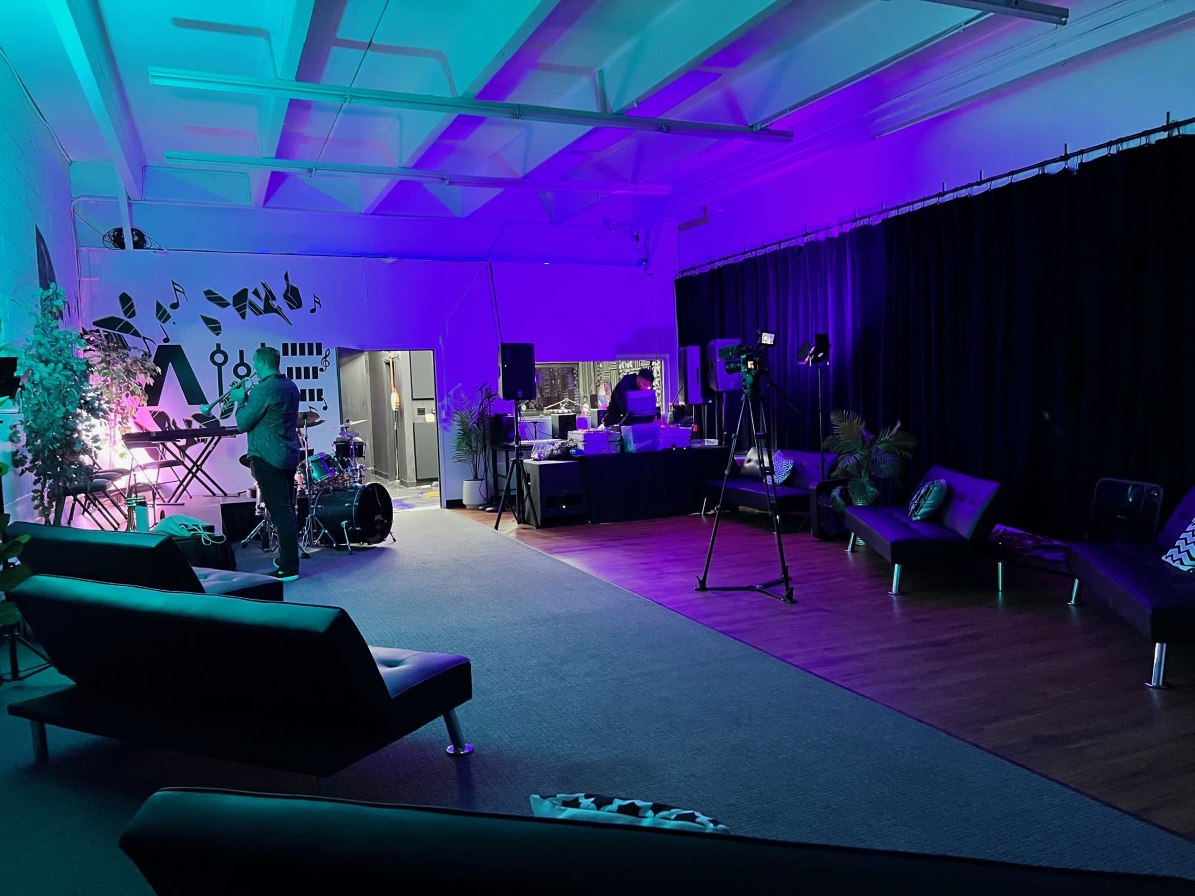 A lively interior space set up for a music event, featuring a stage with instruments, lighting, and seating areas.