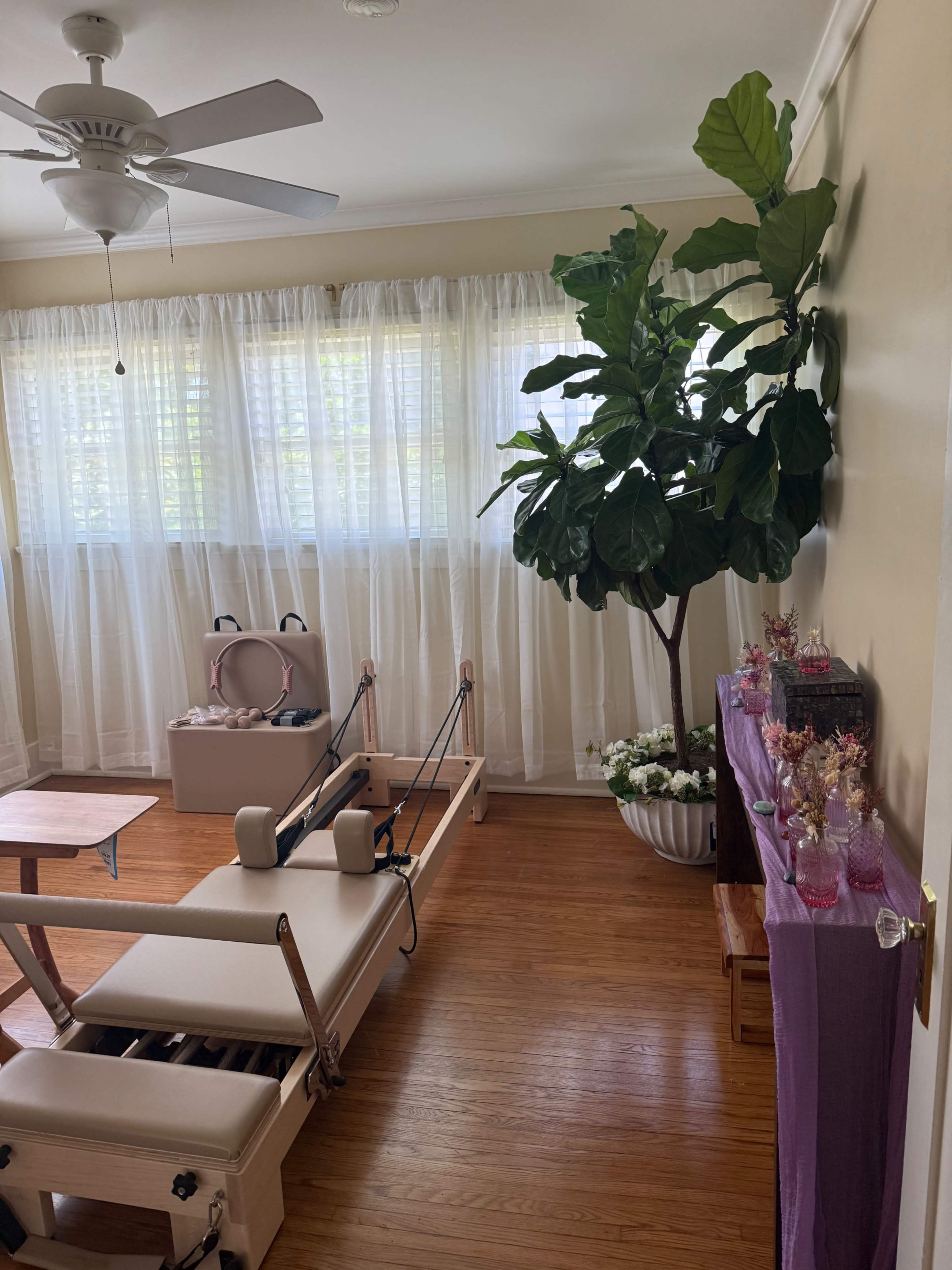 The image shows a bright room with large windows, a reformer Pilates machine, a decorative plant, and a table adorned with various small items.