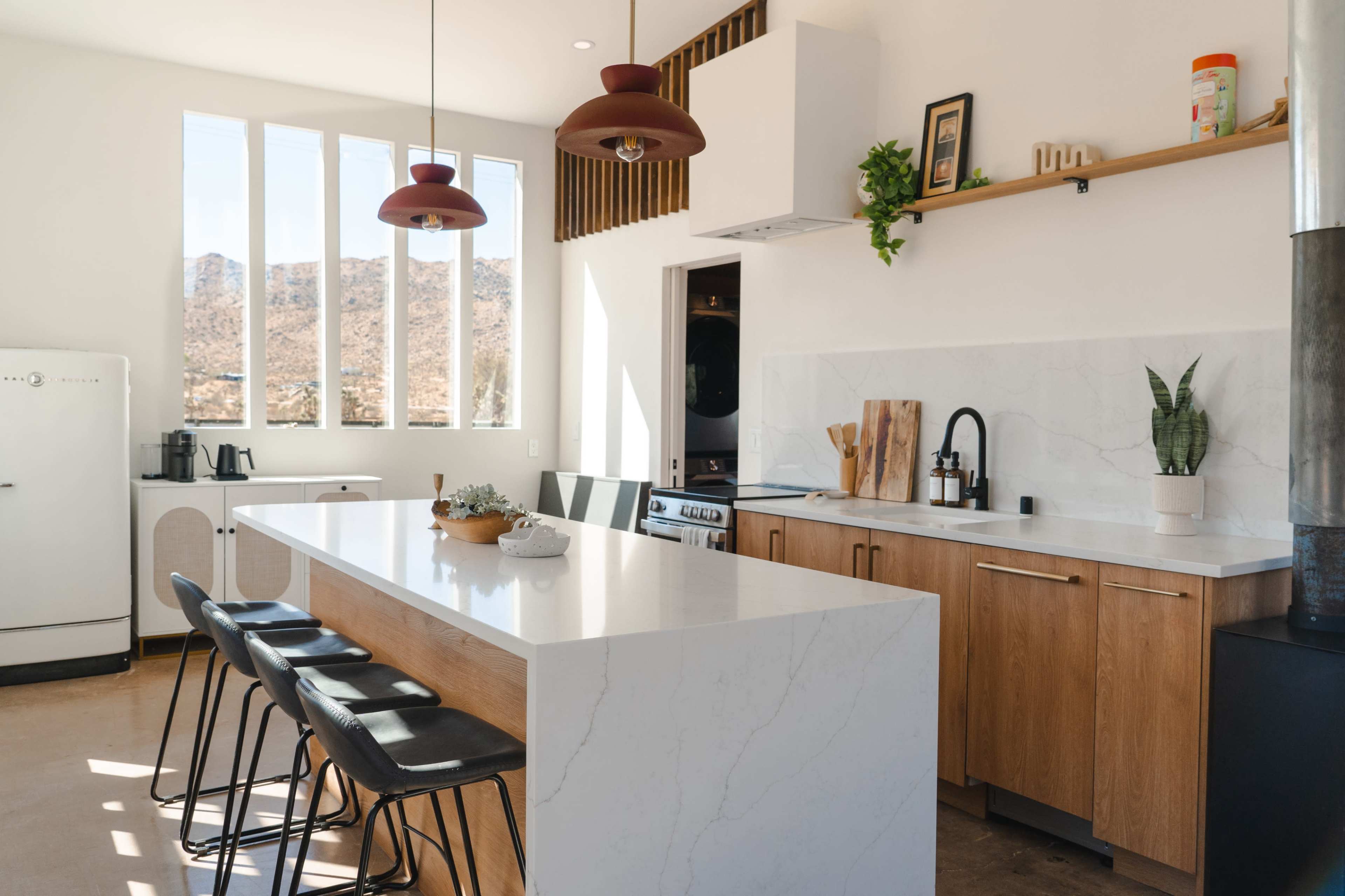 The image shows a modern kitchen with a large white marble island, wooden cabinetry, and large windows that provide views of a rocky landscape.