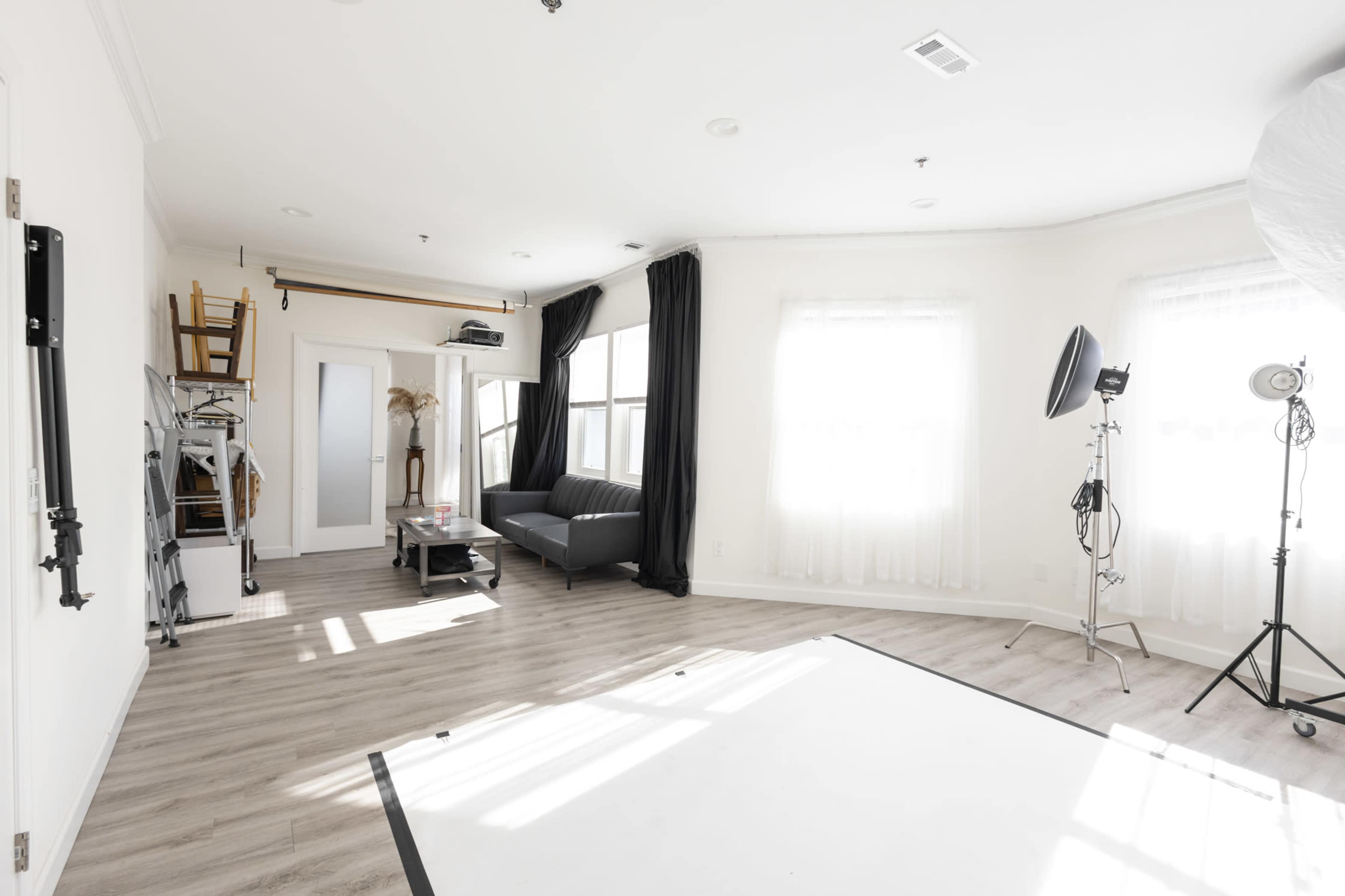 The image shows a bright, spacious photography studio with a sofa, large windows, and lighting equipment.