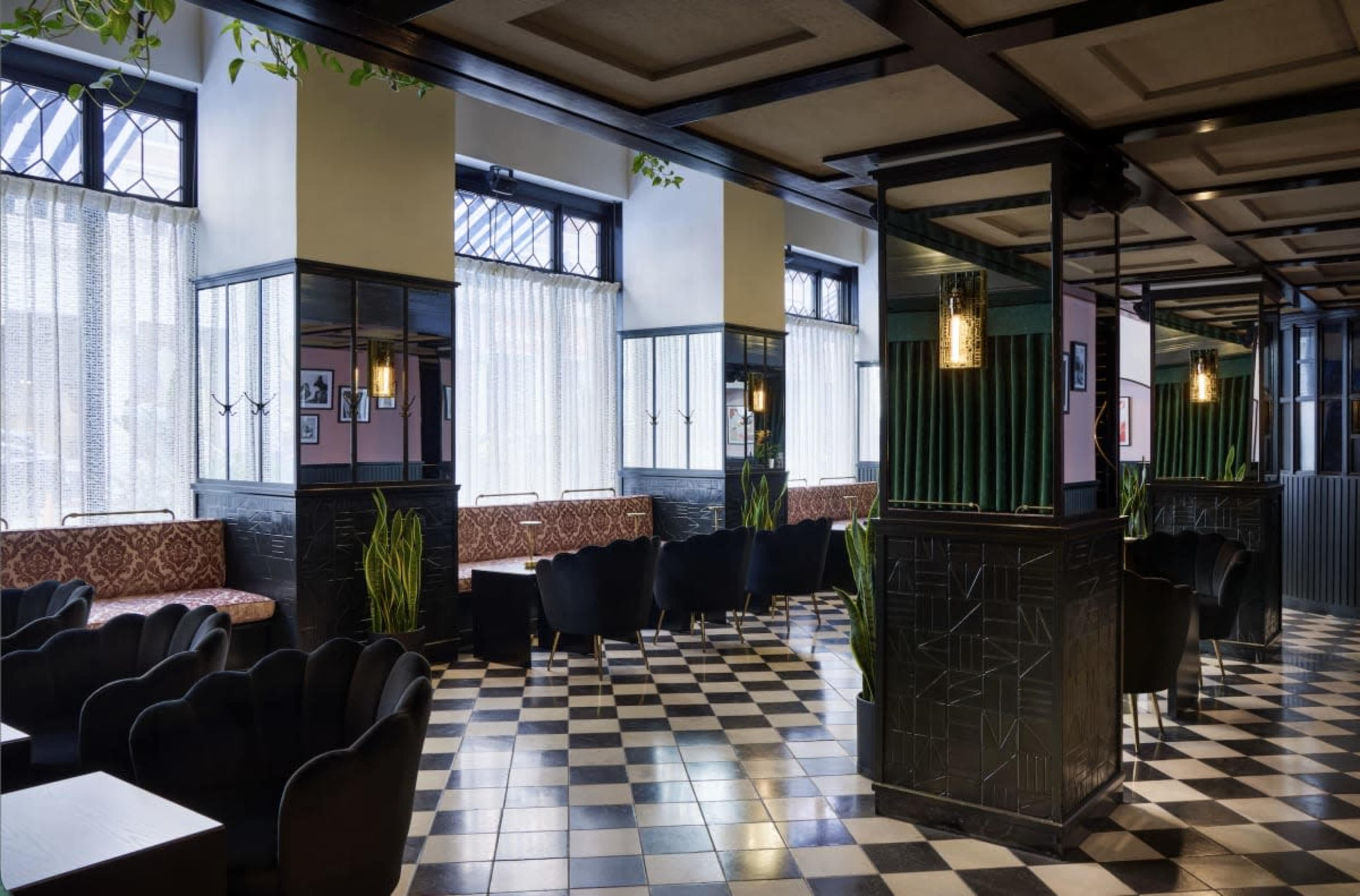 The image shows a modern café interior with patterned black and white flooring, seating areas, and large windows adorned with sheer curtains.