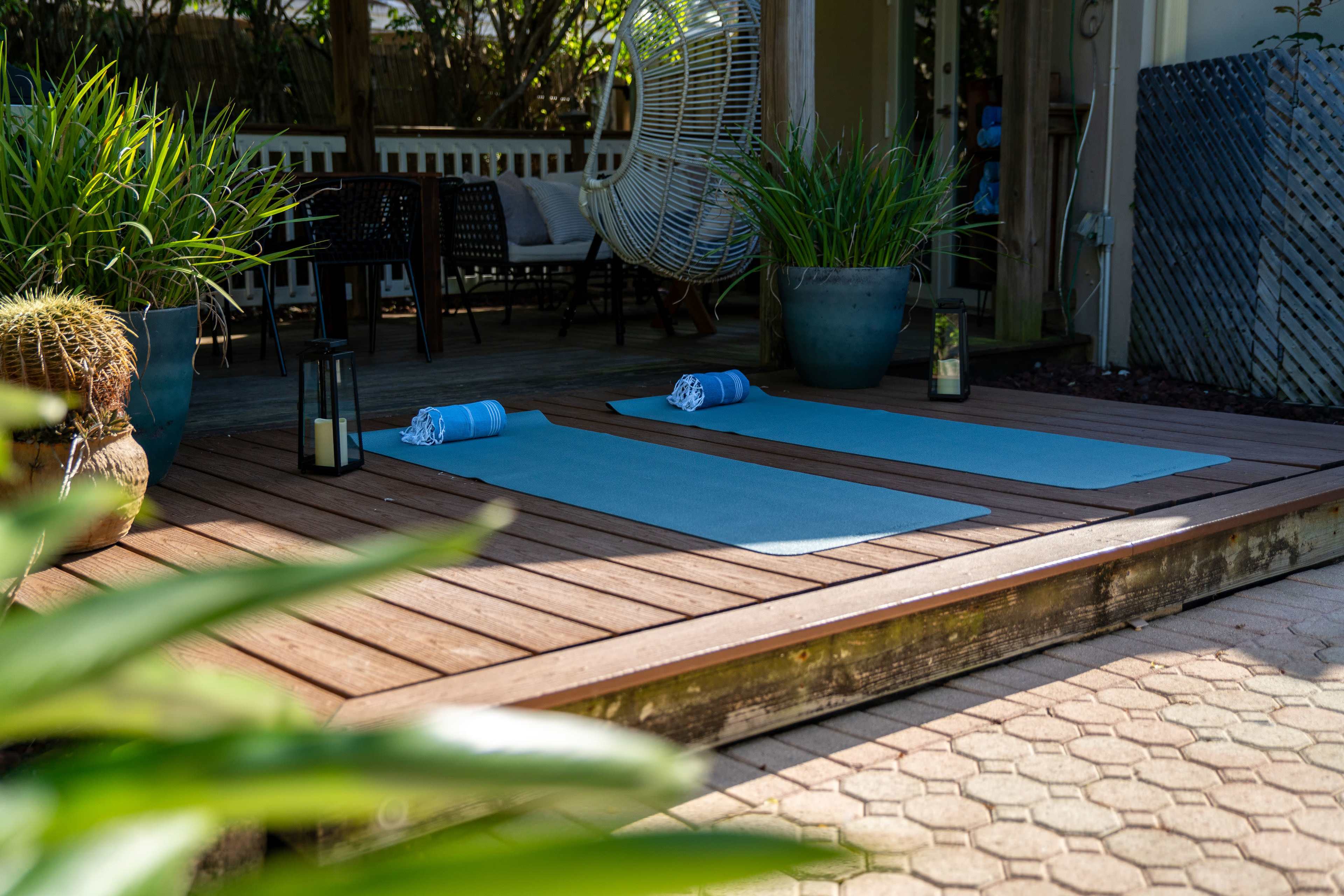 Two yoga mats are laid out on a wooden deck, accompanied by rolled towels and potted plants.