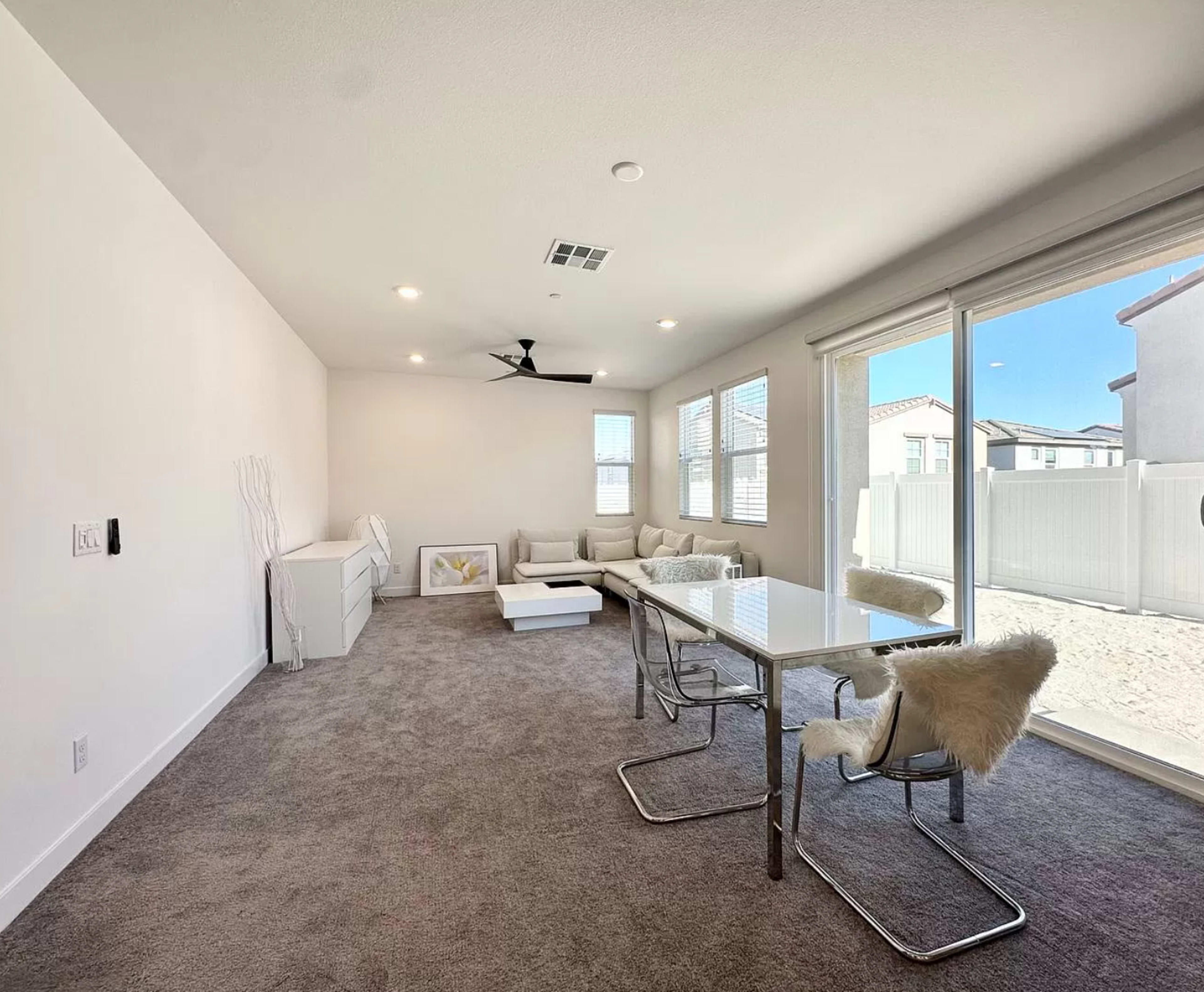 A modern living room features a light-colored couch, a glass-top dining table, and large windows overlooking a fenced outdoor area.