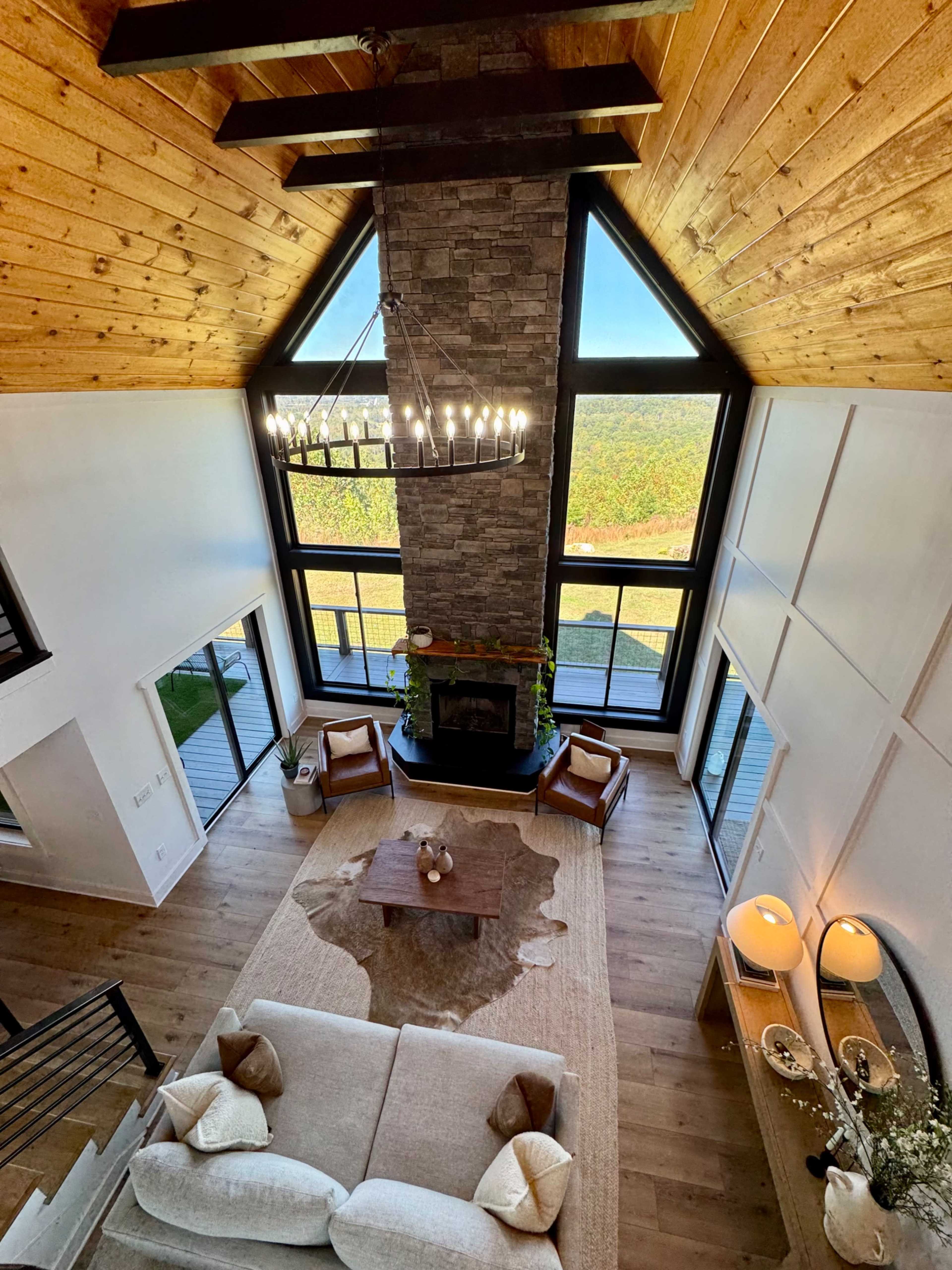 The image shows a spacious living room with large windows, a stone fireplace, a wooden ceiling, and a seating area with a sofa and chairs.