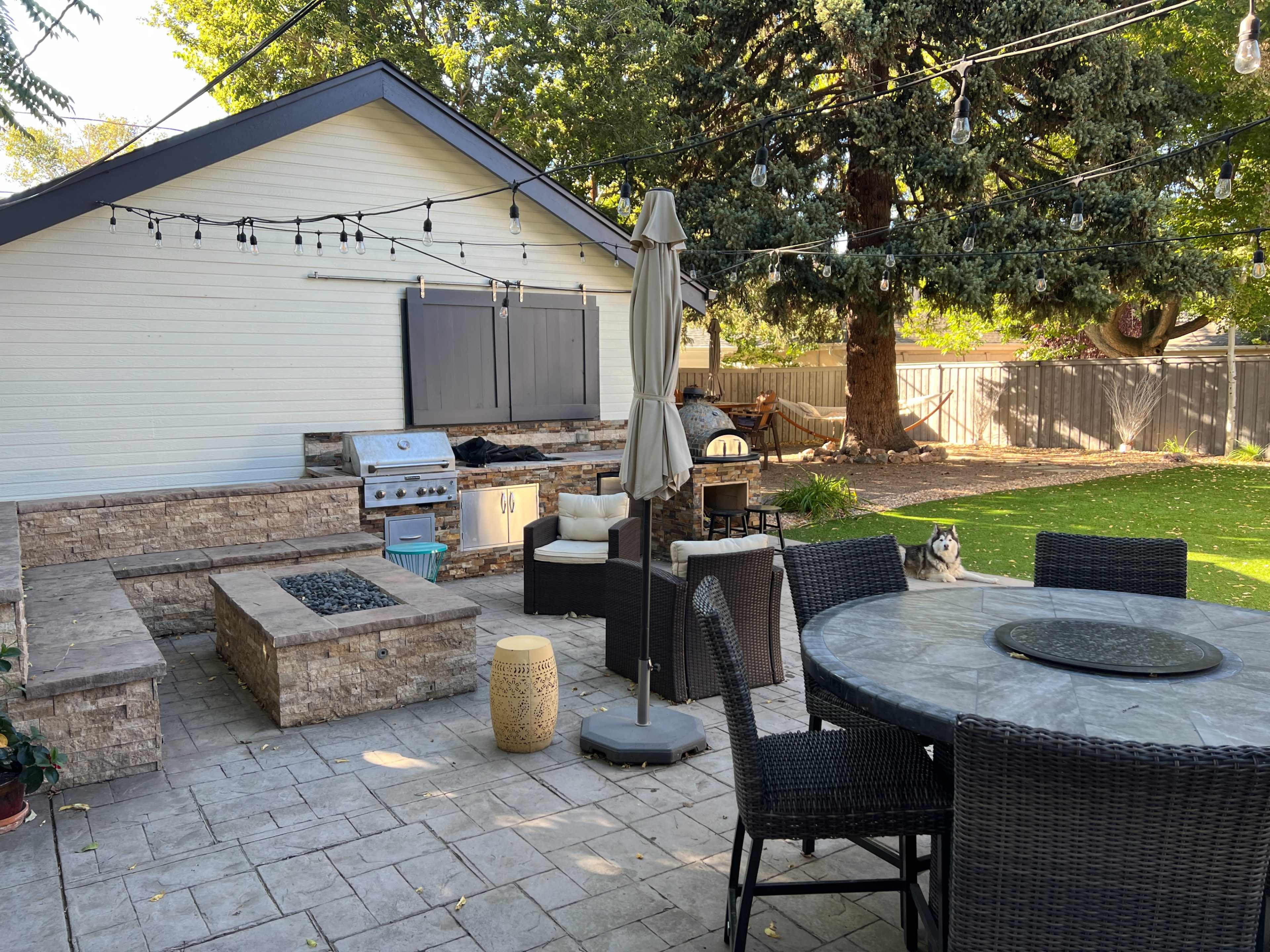 The image shows a backyard patio area featuring a stone seating arrangement, a fire pit, a dining table, and a barbecue grill near a house surrounded by trees and string lights.