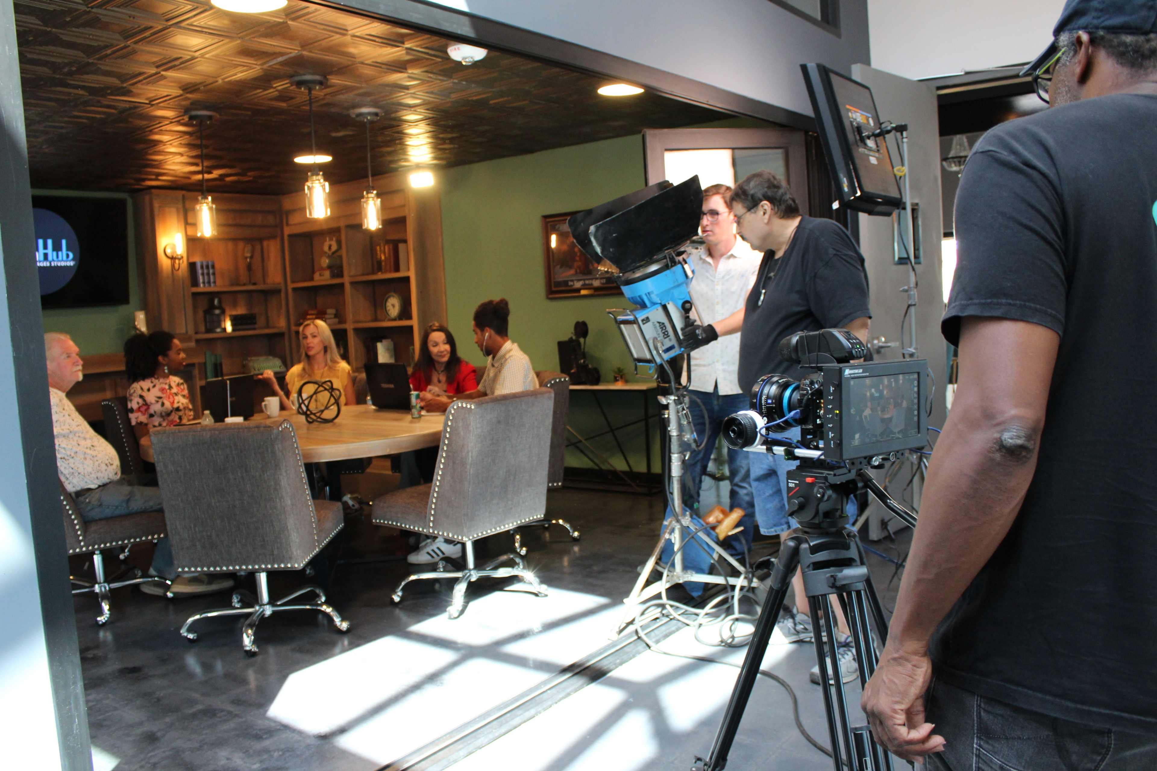A film crew is setting up equipment in a meeting room where a group of people is seated around a table.