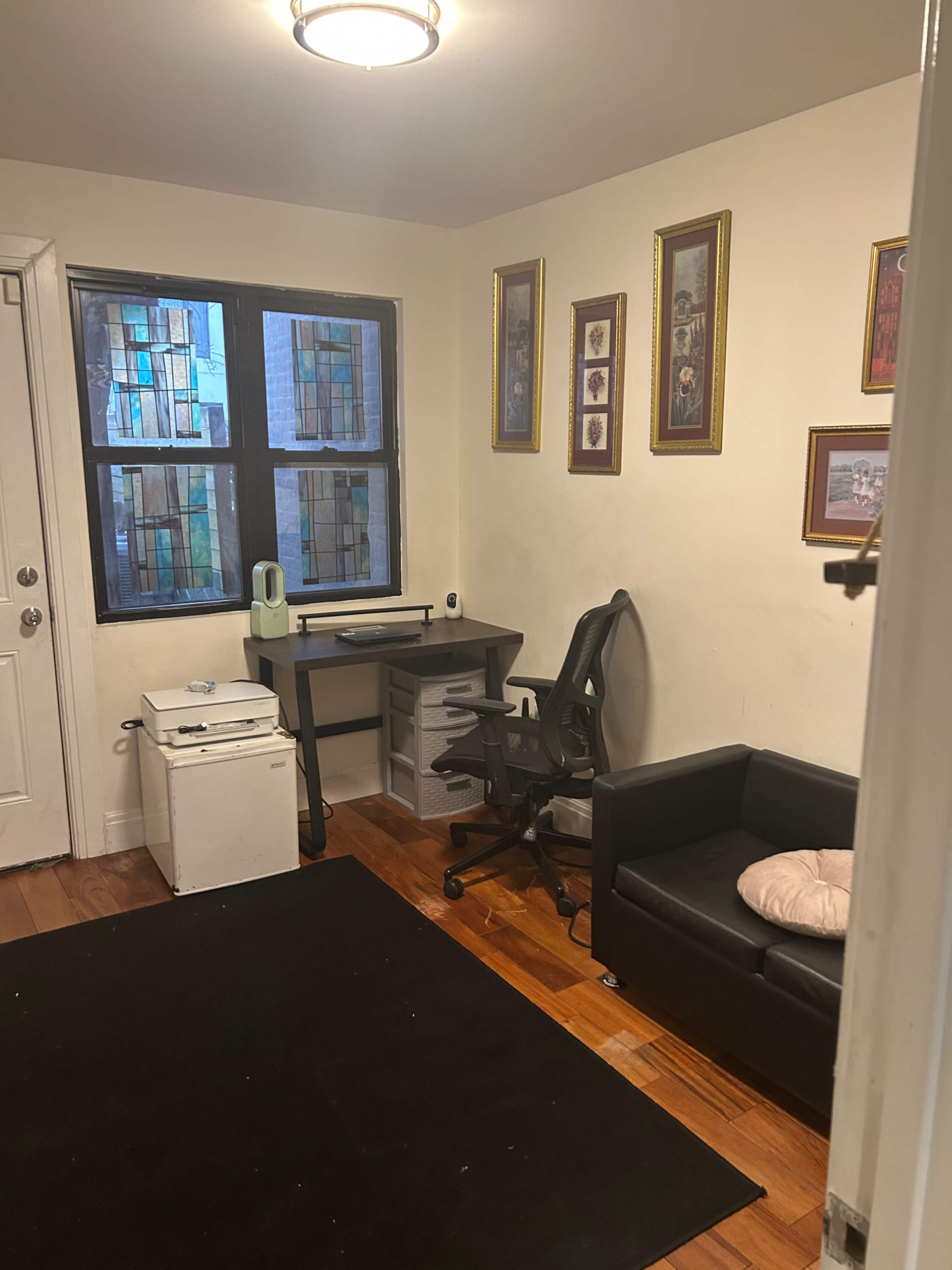 The image shows a small room with a black rug, a desk with a chair, a white compact filing cabinet, and a black couch against the wall, featuring framed artwork and windows with patterned glass.
