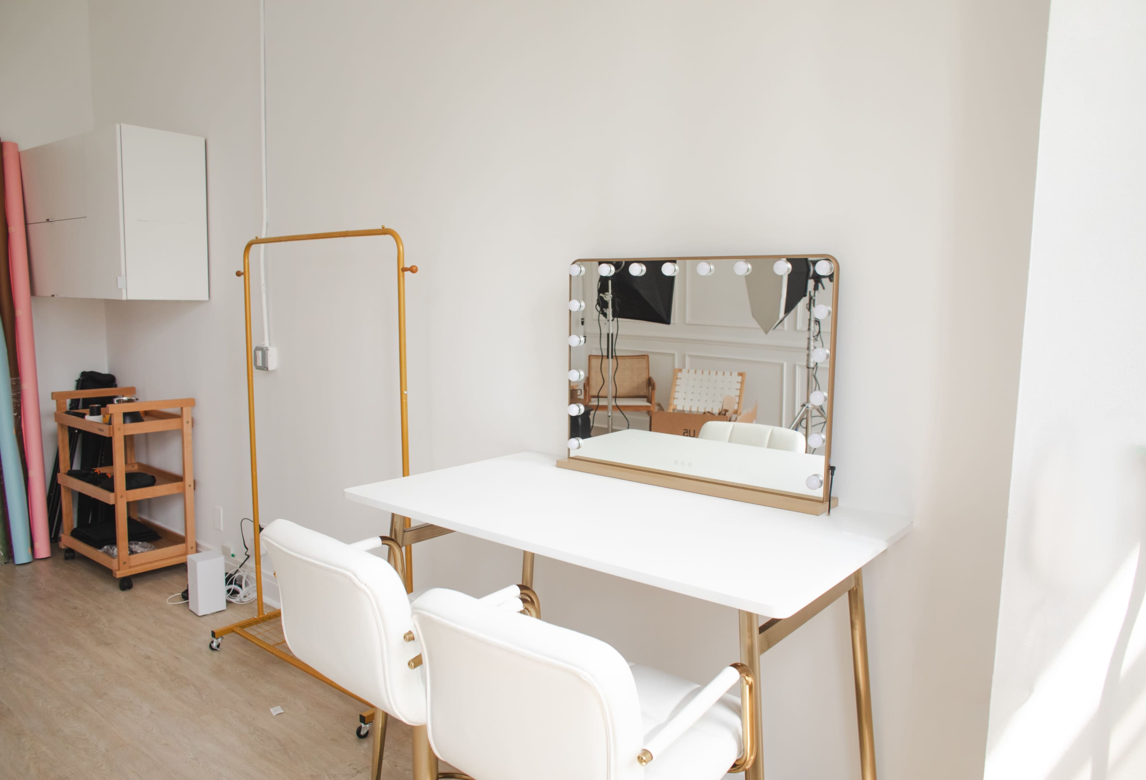 The image shows a well-lit dressing area featuring a white table with two gold-accented chairs and a large mirror framed with lights.