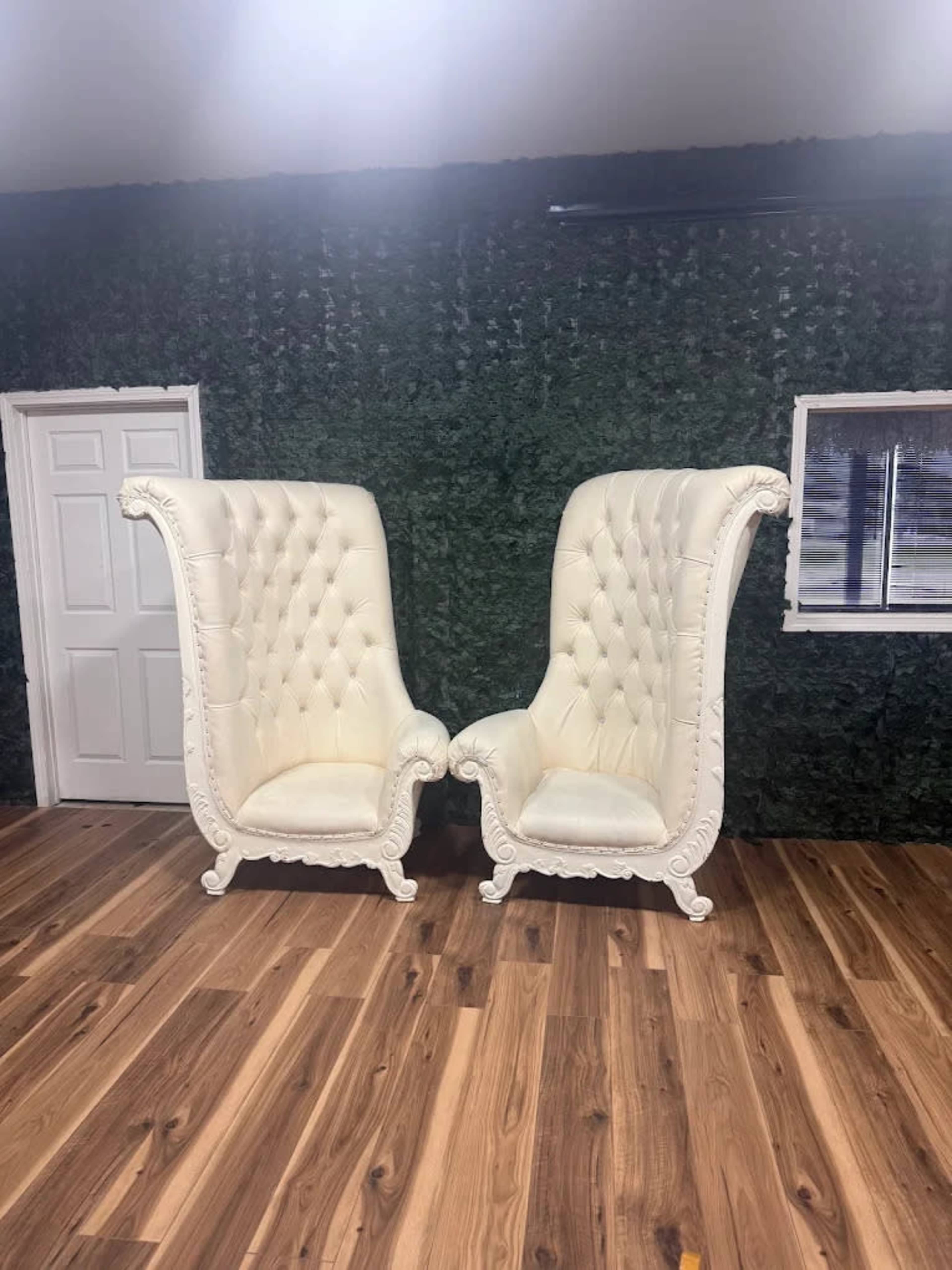 Two ornate white high-backed chairs with tufted upholstery are positioned facing each other on a wooden floor, against a backdrop of a green wall and a door.