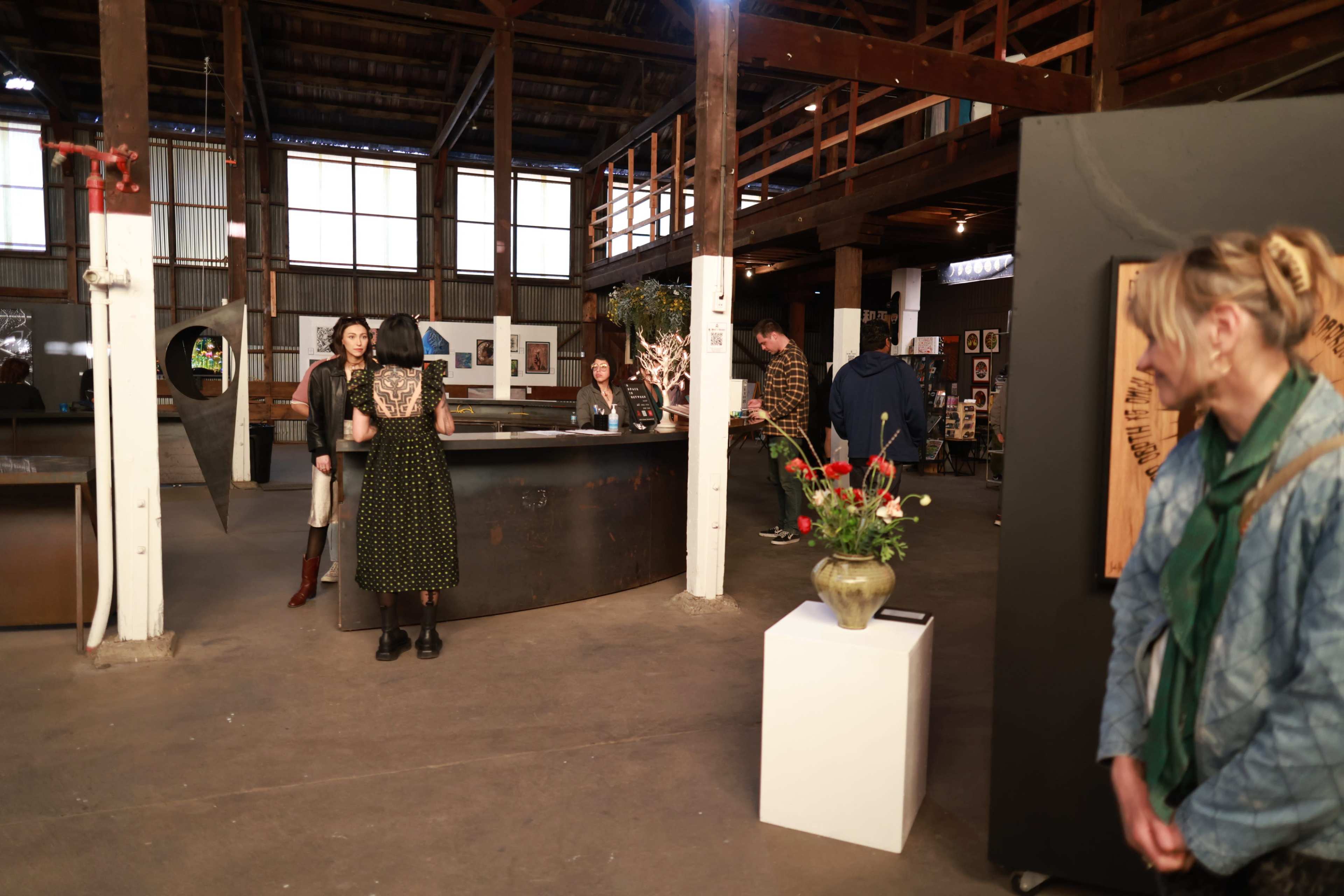 The image shows an art exhibition in a spacious industrial venue, with several attendees interacting and viewing various artworks displayed.