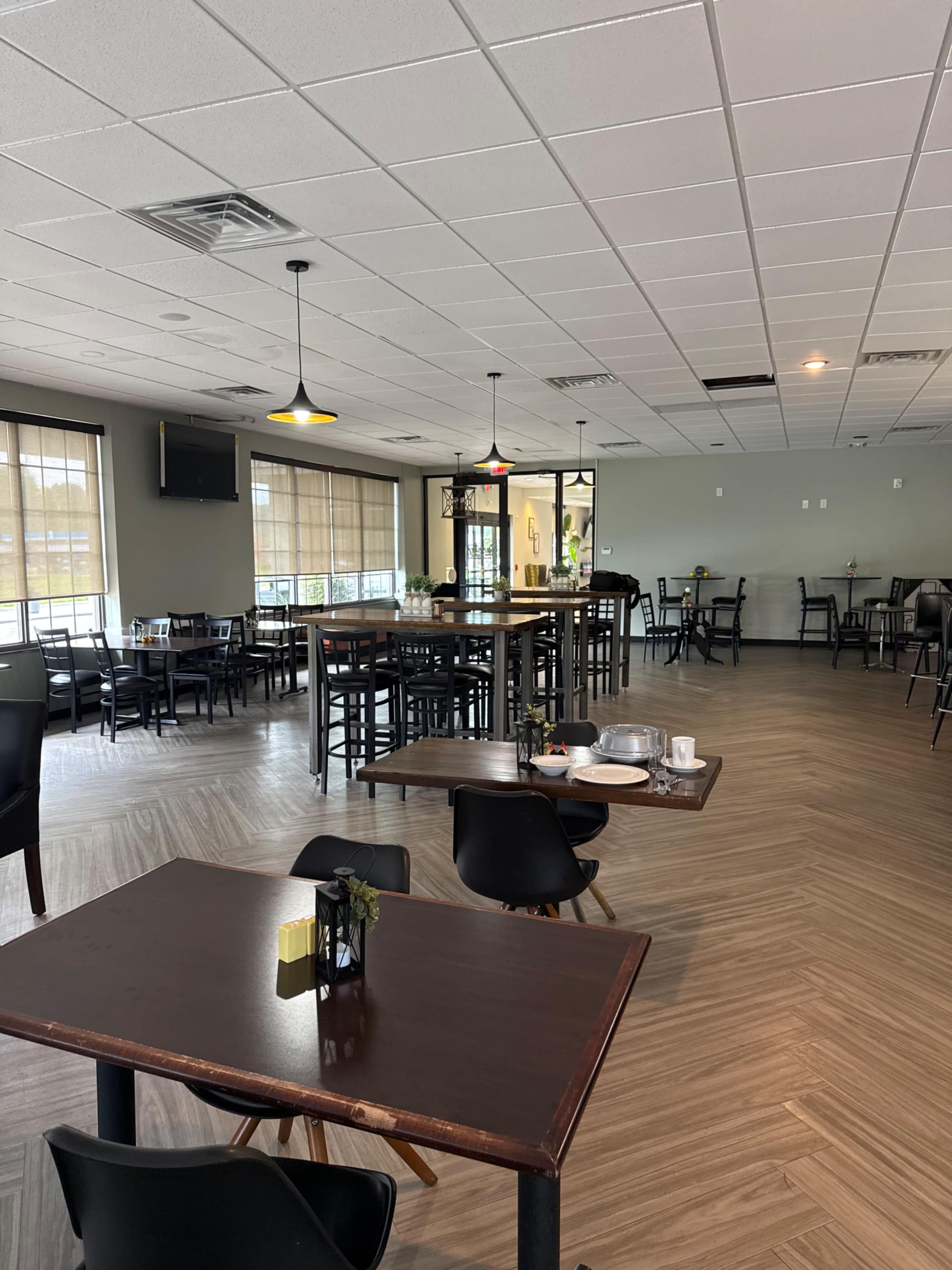 The image shows a spacious restaurant interior with several tables and chairs arranged neatly, large windows allowing natural light, and a television mounted on the wall.