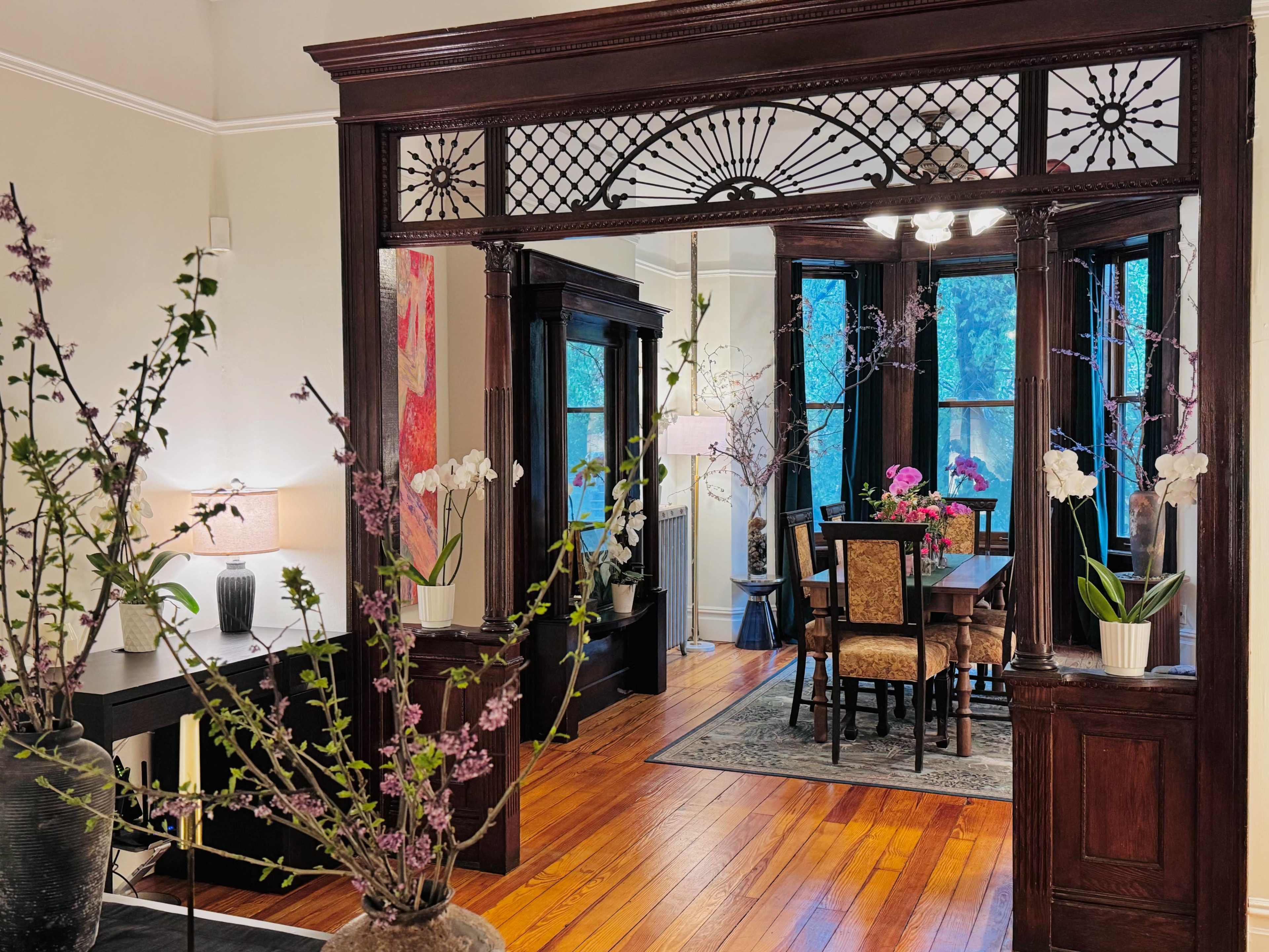 The image shows a well-furnished dining area with a wooden table surrounded by chairs, framed by ornate wooden architecture and large windows letting in natural light.